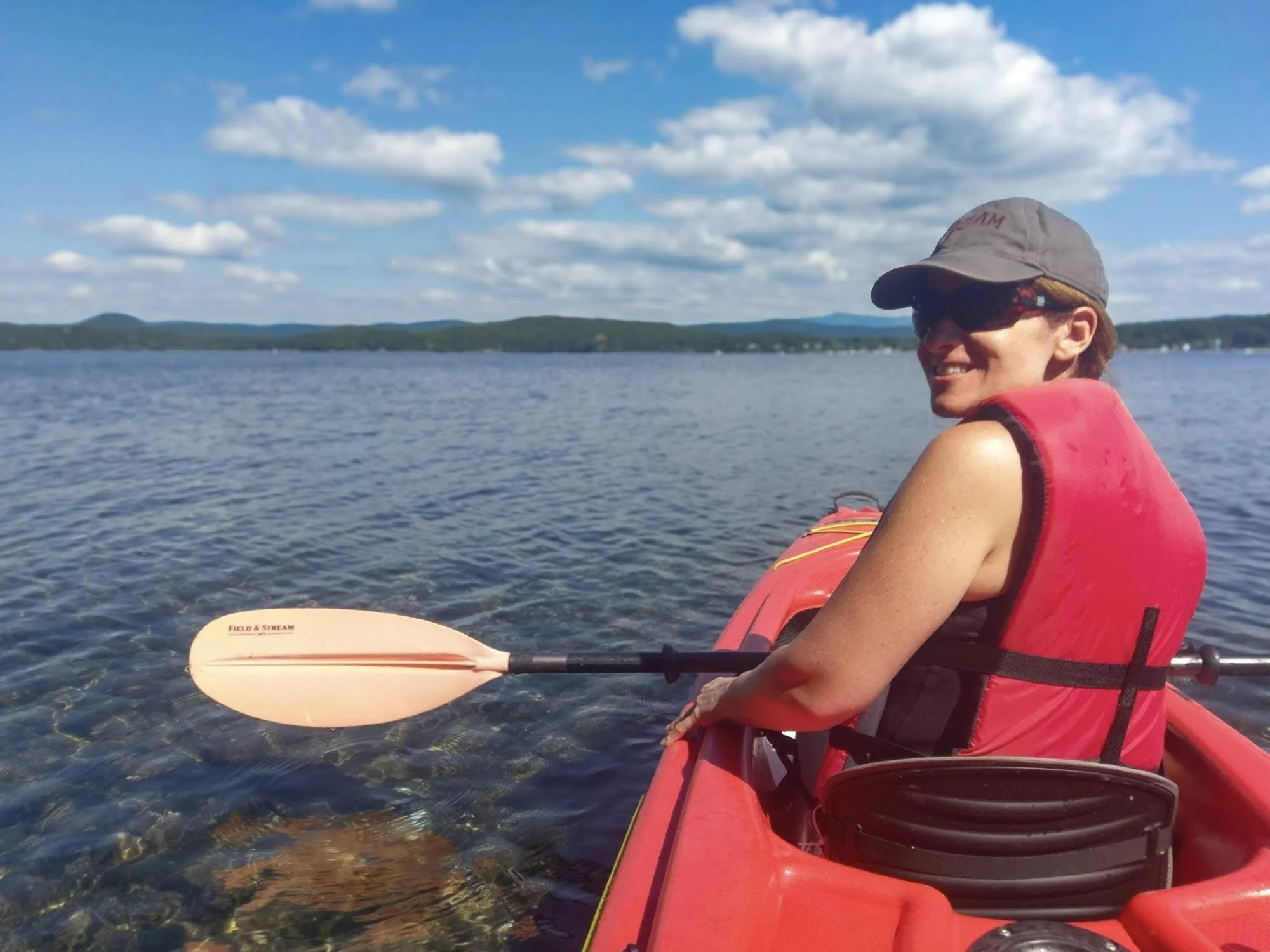 Woman in a red life jacket and gray cap kayaking on a lake with a scenic view of distant hills and a blue sky with clouds.