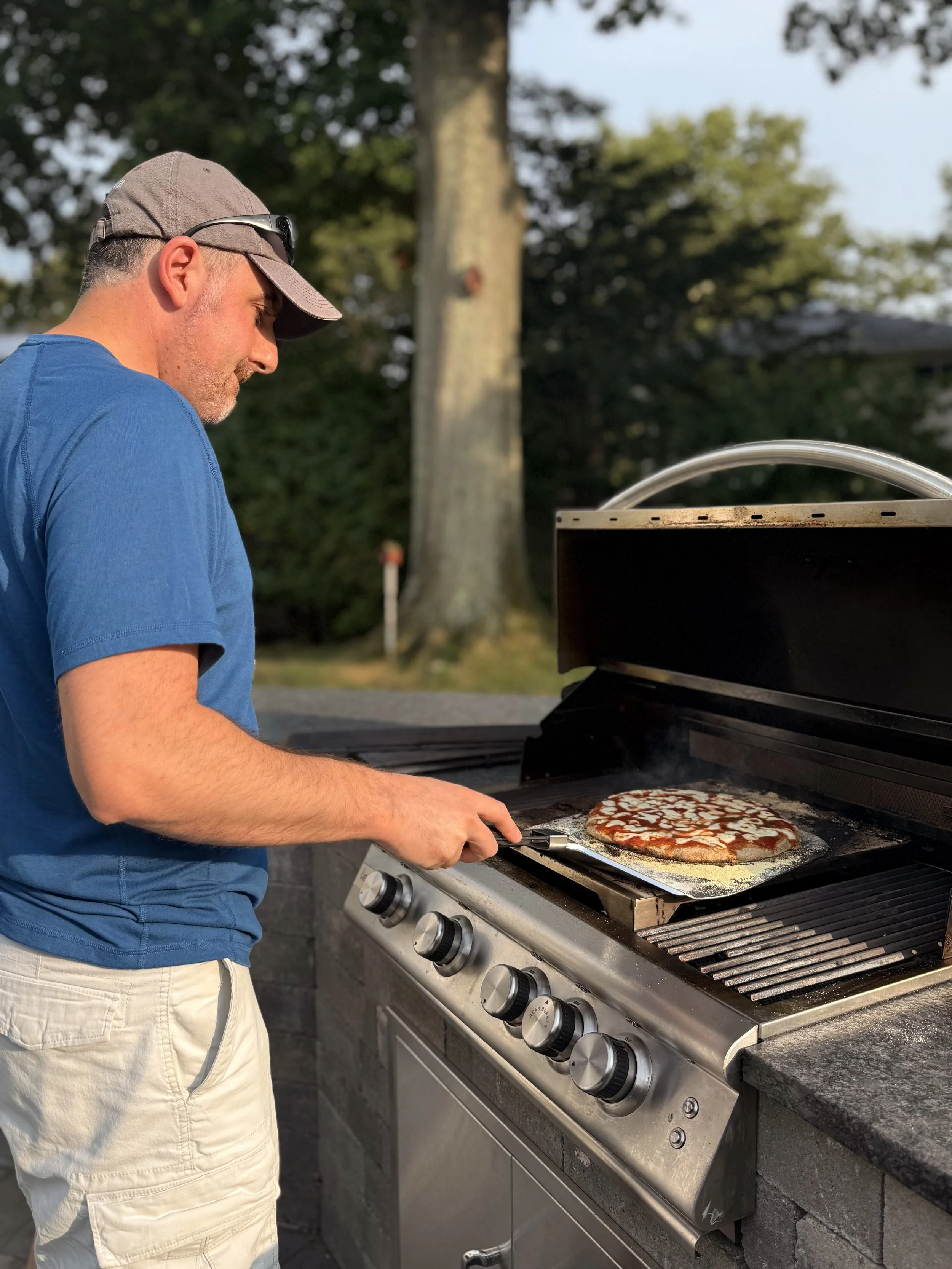 Man in a blue shirt and beige shorts is grilling a pizza on a barbecue grill outdoors during daytime. The pizza has cheese and tomato toppings.