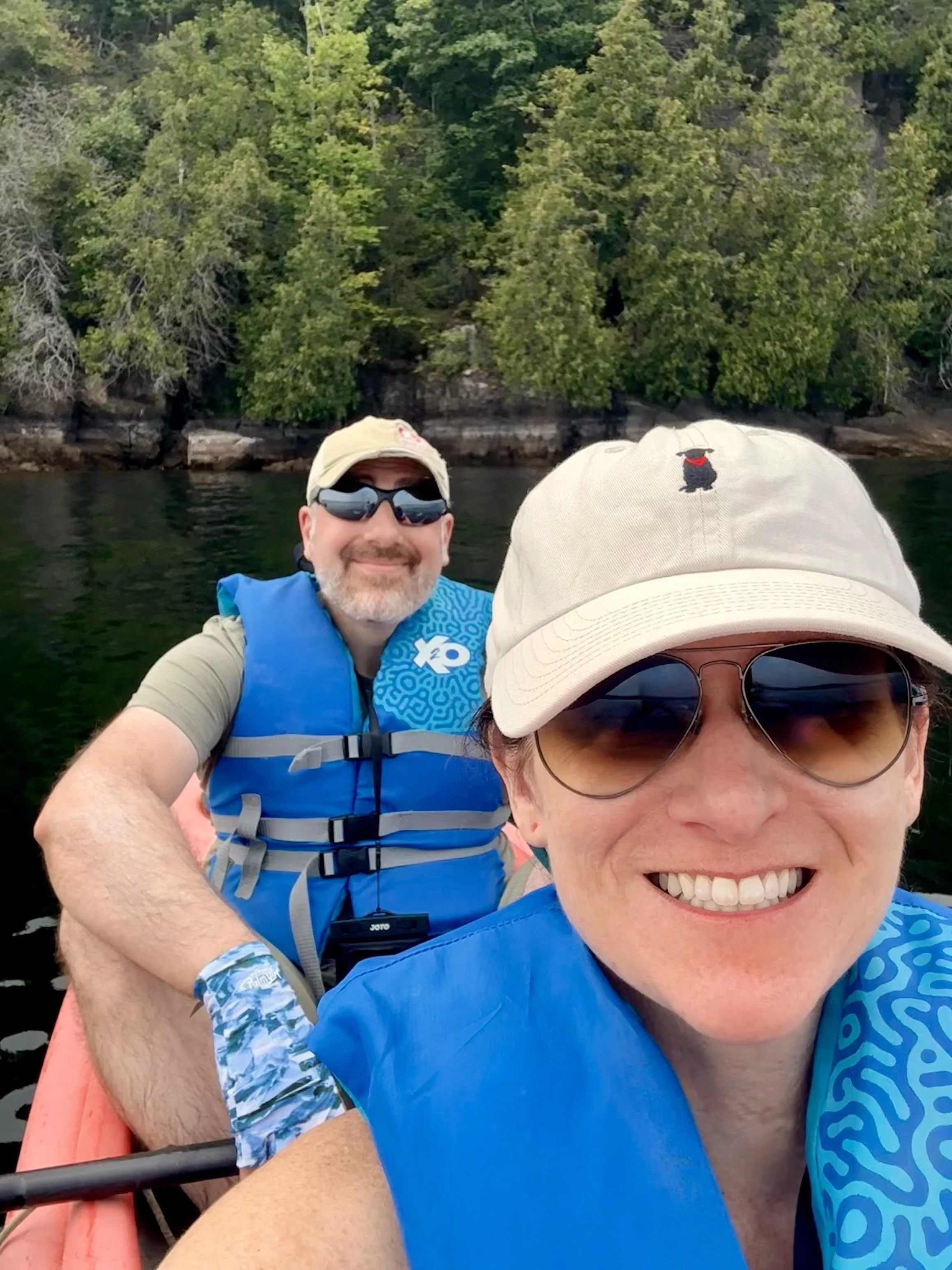 A smiling woman and man sitting in a kayak on a body of water, with a background of green trees and rocks. They are wearing sunglasses, hats, and life jackets.