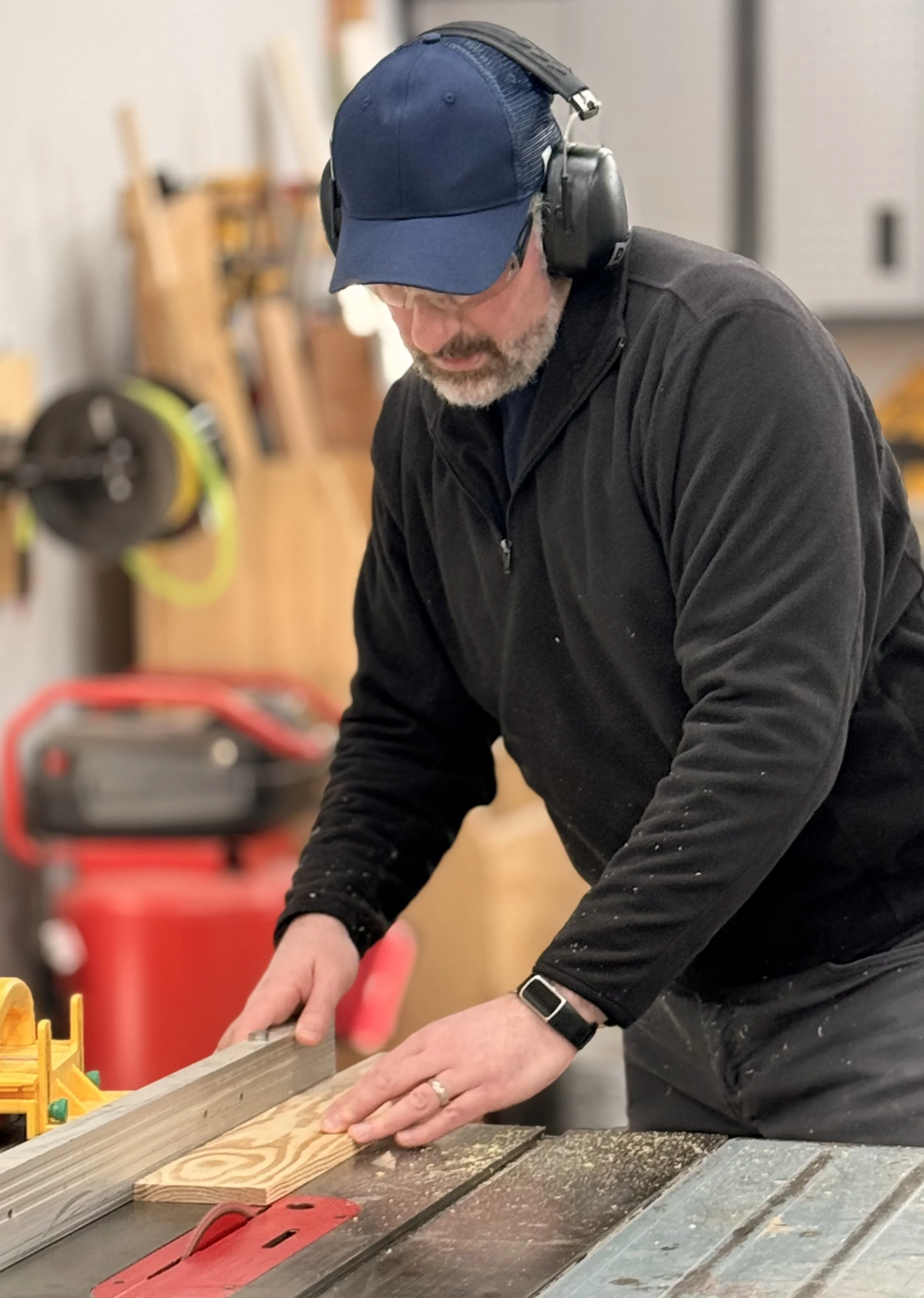 A man in a blue baseball cap, black jacket, and safety earmuffs is working in a woodworking shop, using a sled on a table saw to cut a piece of wood.