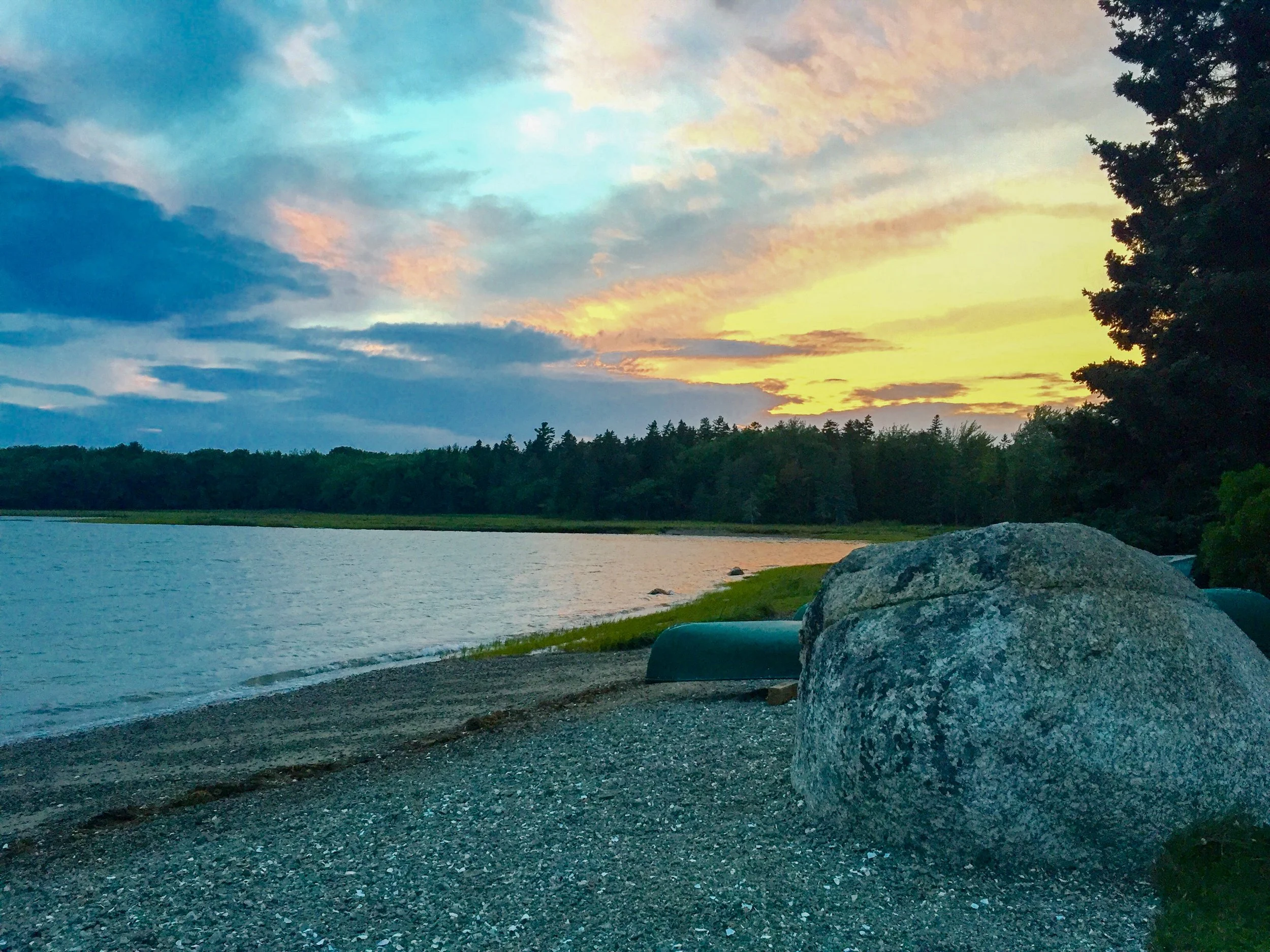 Scenic view of a lake at sunset with colorful sky and clouds, surrounded by trees, with a large rock and small boats on the shore in the foreground.