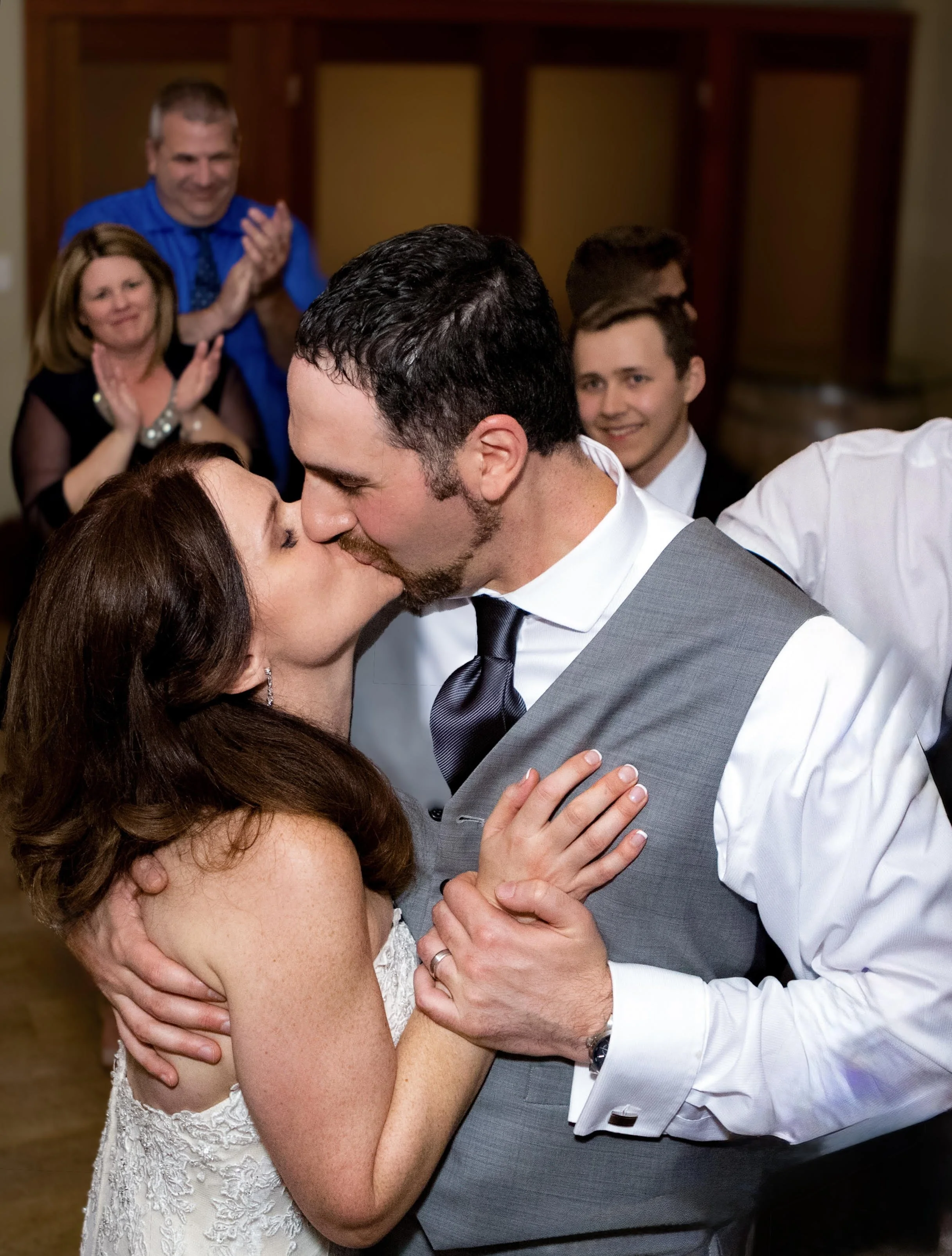 A couple sharing a kiss during their wedding dance, surrounded by happy guests clapping and smiling in the background.