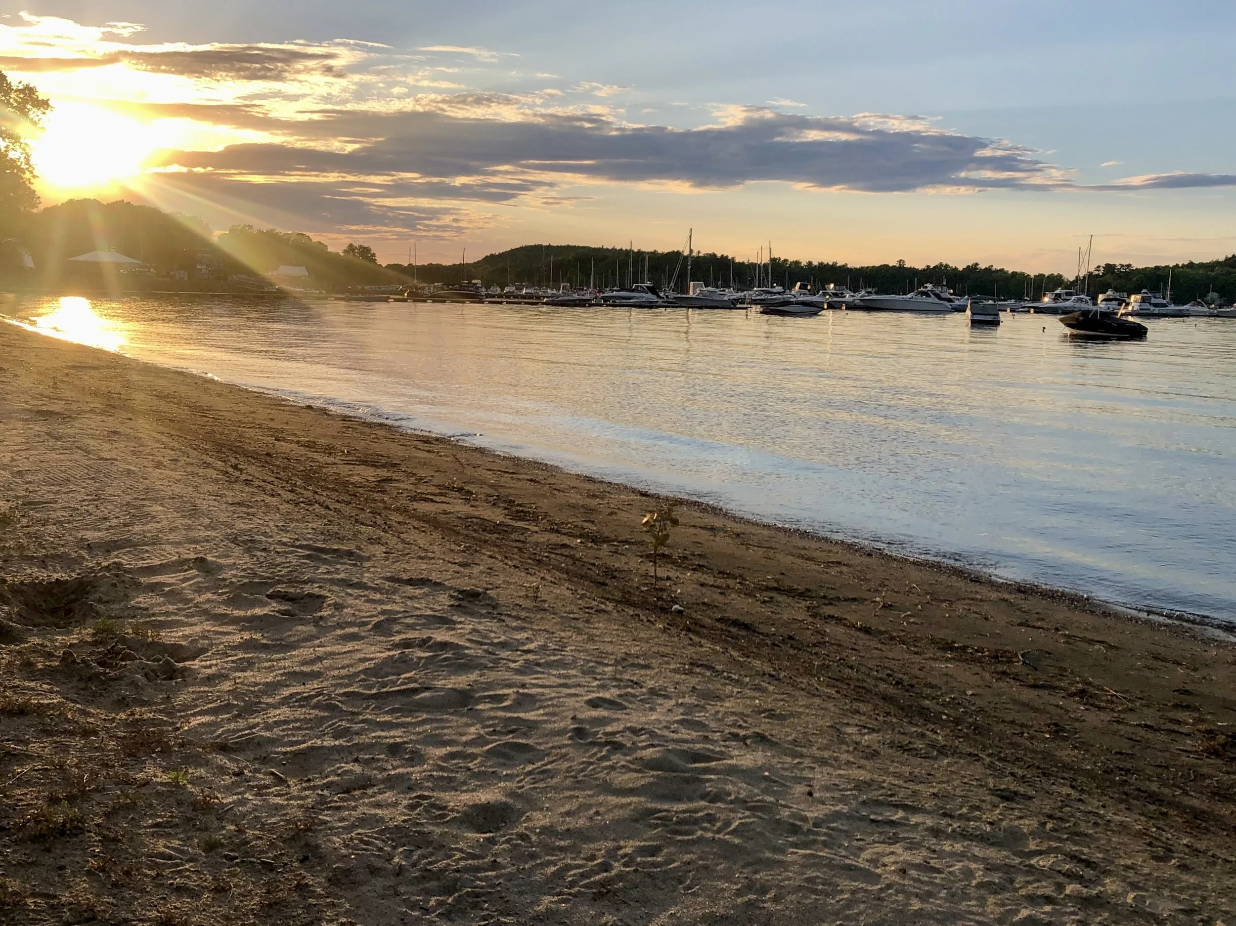 Sunset over a marina with boats docked on the water, viewed from a sandy beach with footprints.