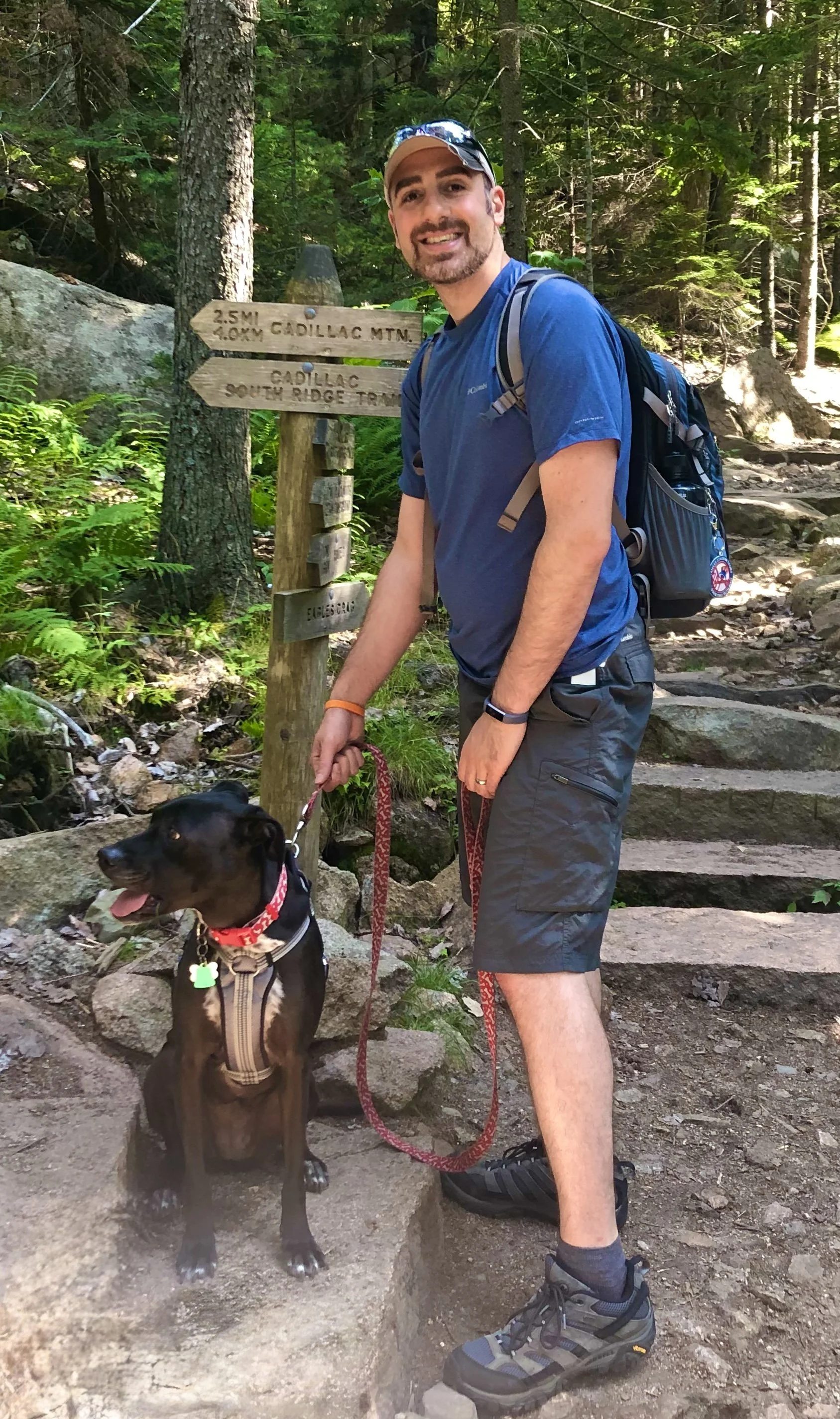 A man wearing outdoor clothing and a backpack holding a dog on a trail in a forest with a wooden signpost in the background.