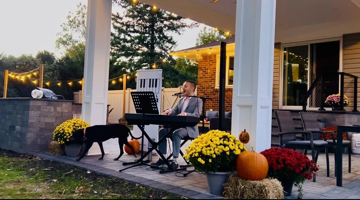 A man in a suit playing a keyboard on a decorated outdoor porch during evening, with pumpkins, flowers, and hanging lights. Fall backyard concert.