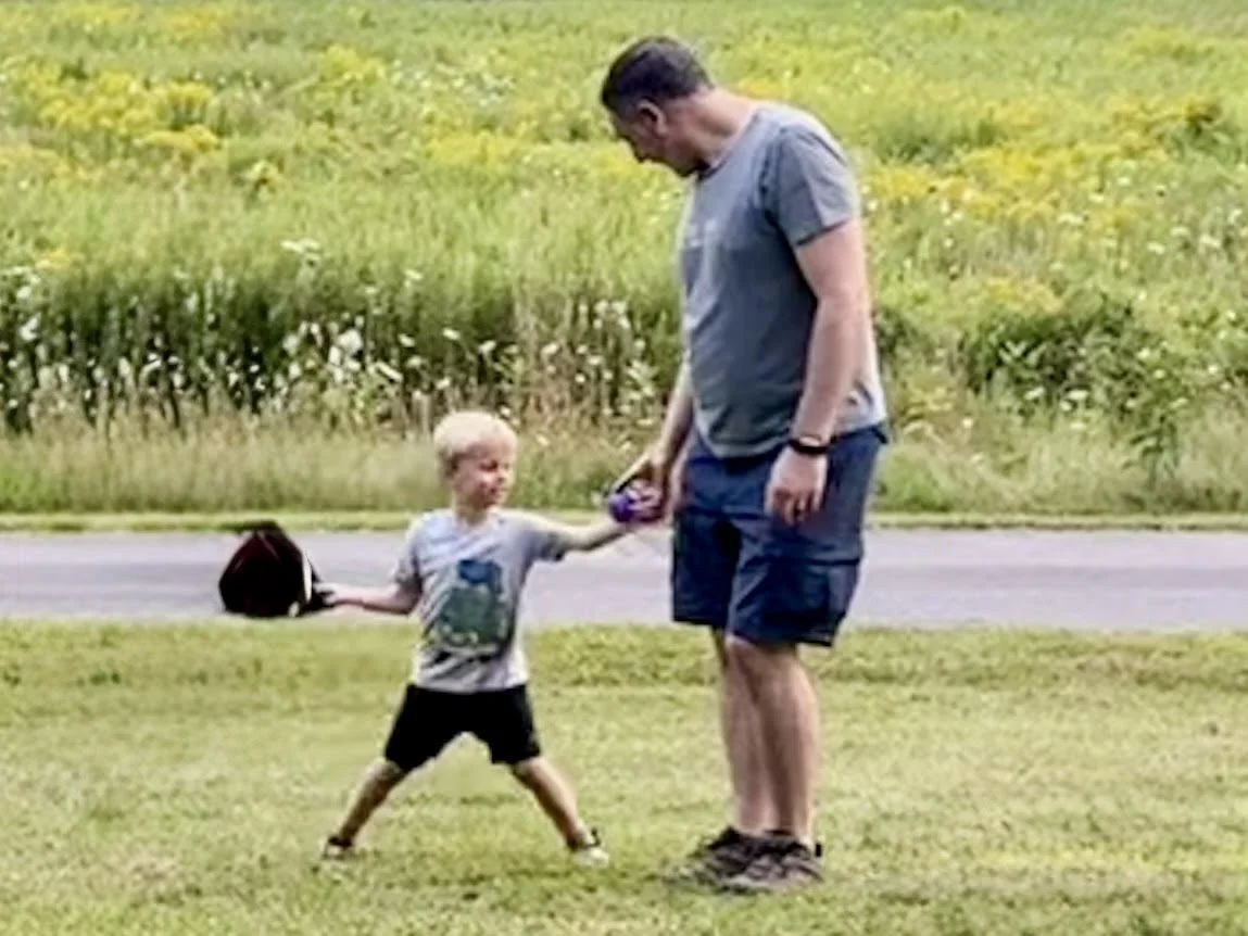 A young boy handing an adult man a baseball in a grassy field. They are having fun being outdoors and playing baseball.