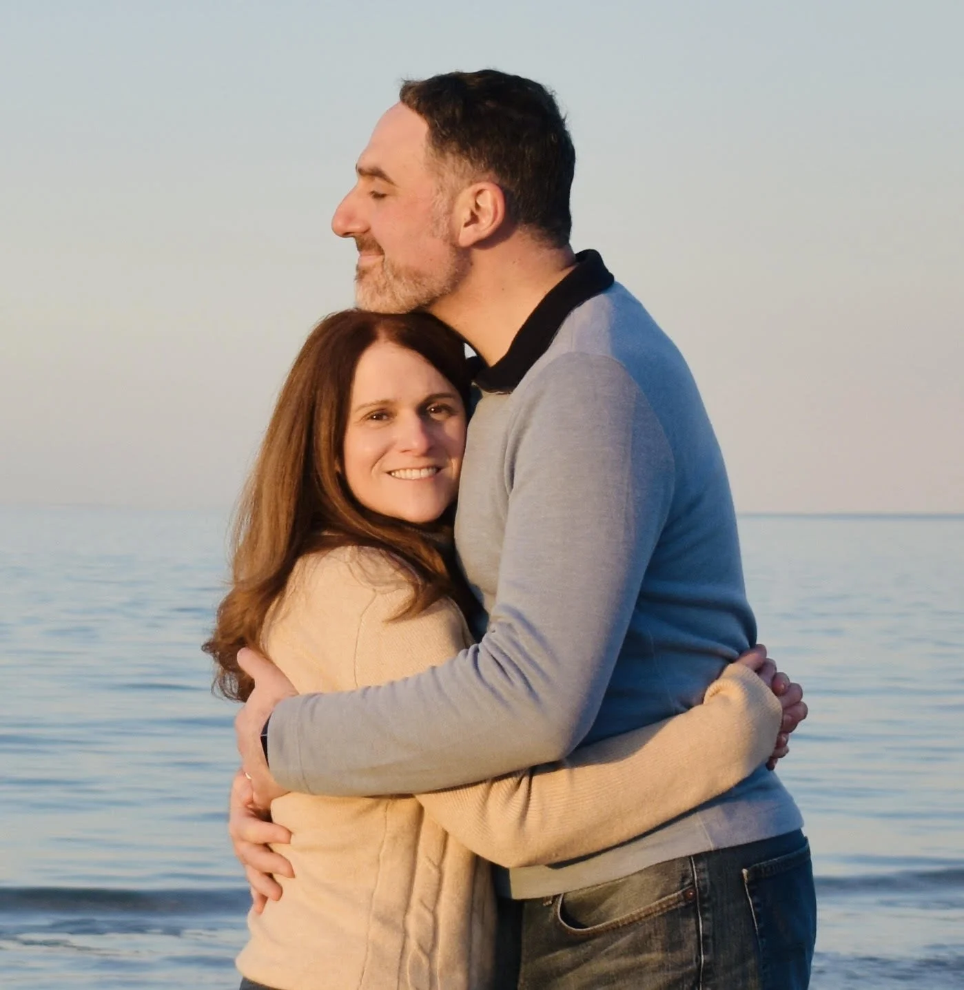 A couple hugging by the water, with clear skies in the background. The woman has long brown hair and is smiling at the camera. The man has short dark hair and a beard, with his eyes closed and a content expression.