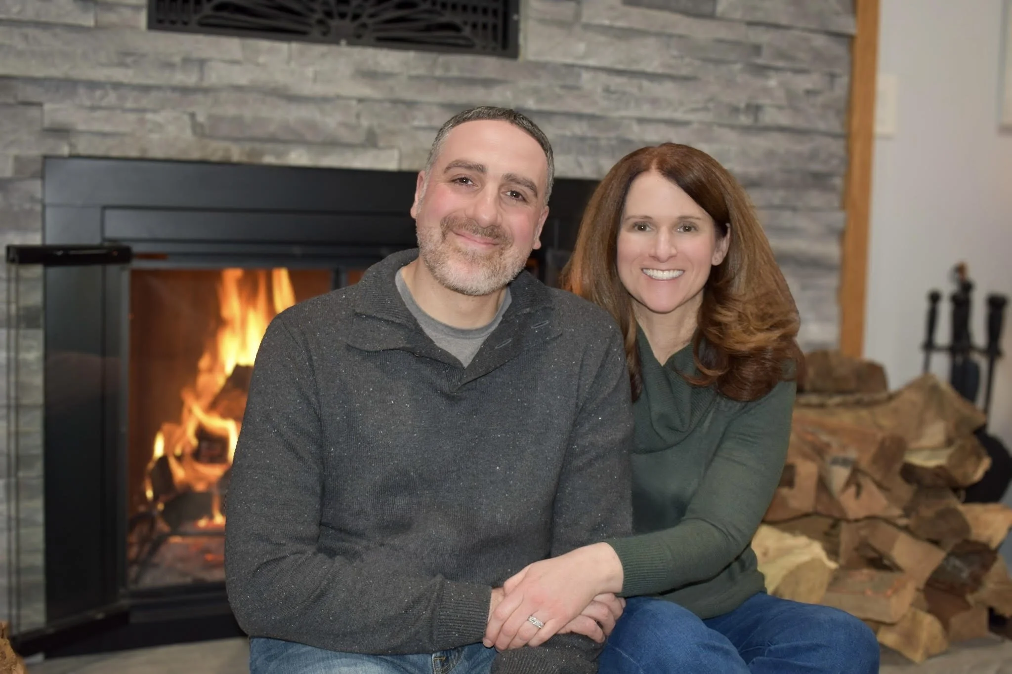 A smiling couple sitting close together in front of a lit fireplace with stacked logs nearby.