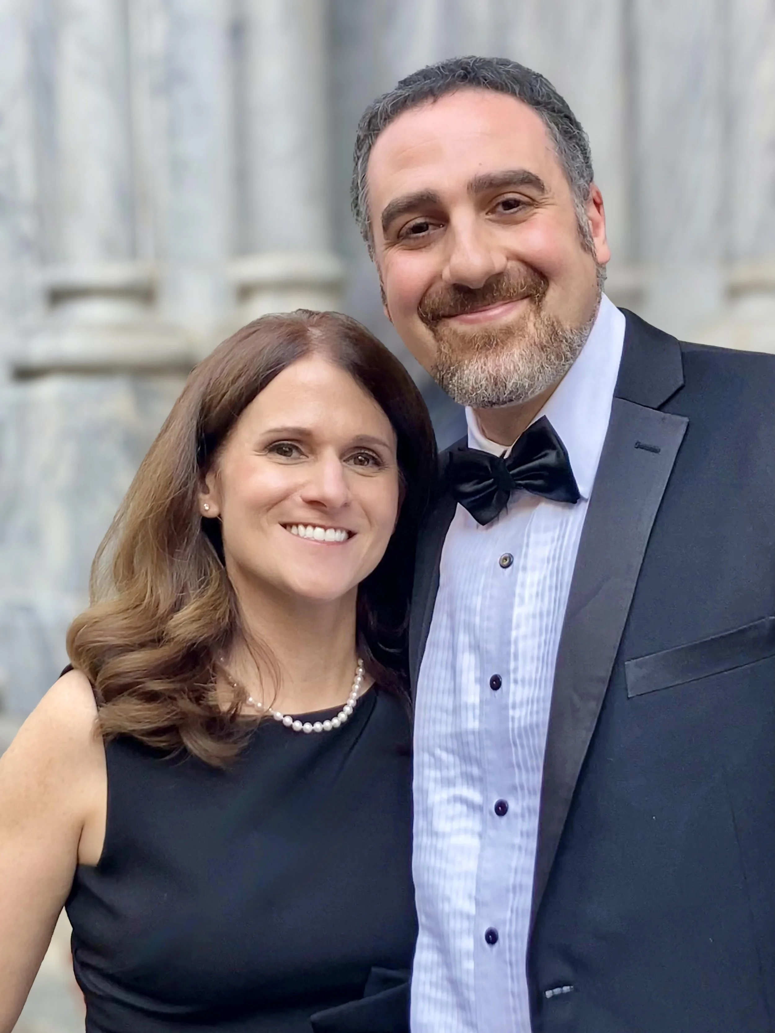 A smiling woman with brown hair in loose curls wearing a black dress and pearl necklace standing next to a smiling man with gray hair and beard, dressed in a black tuxedo with a bow tie, in front of a stone building with columns.