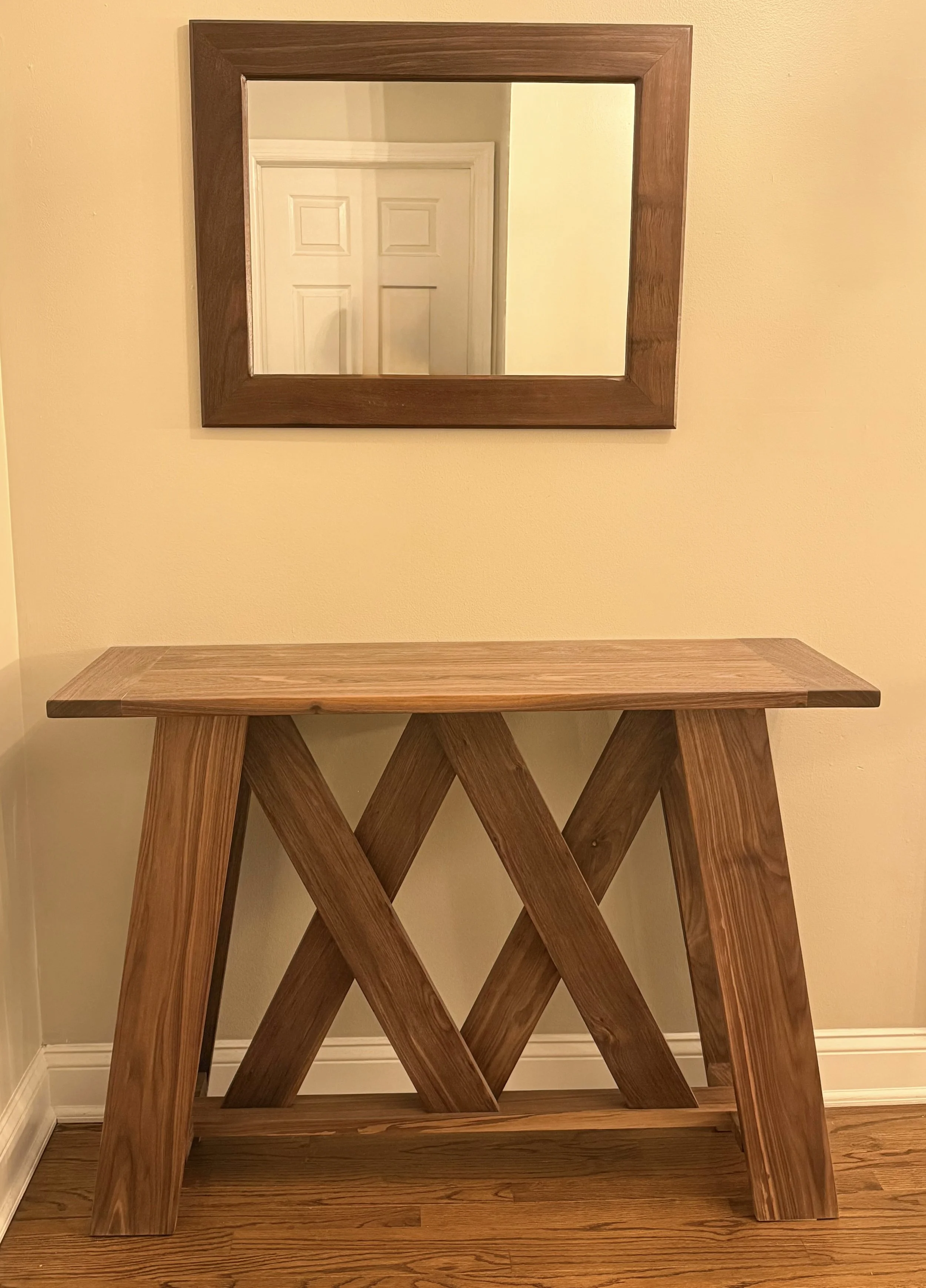 A wooden console table with a cross-braced design underneath, positioned against a beige wall with a rectangular mirror hanging above it.