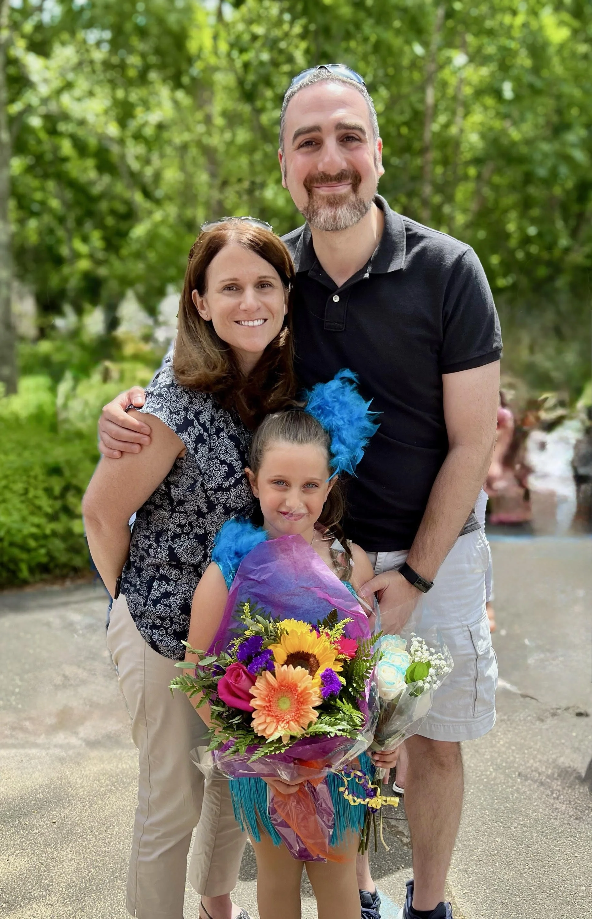 A family of three posing outdoors: a woman, a man, and a girl. The girl is holding a colorful bouquet of flowers.