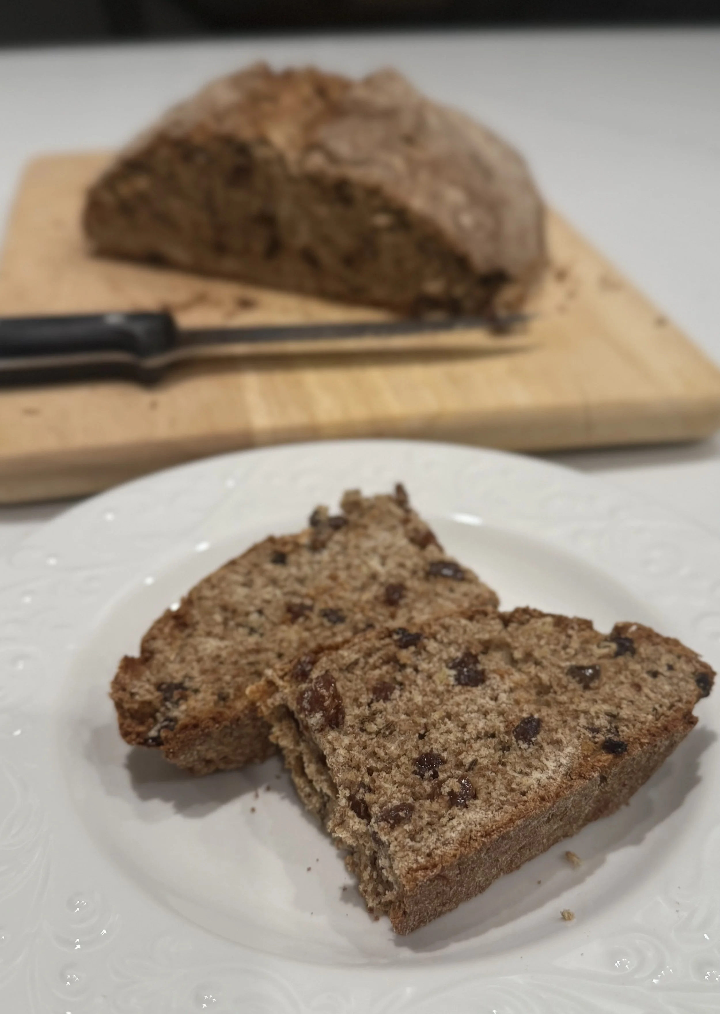 Two slices of Irish soda bread on a white plate with a loaf of Irish soda bread in the background on a wooden cutting board.