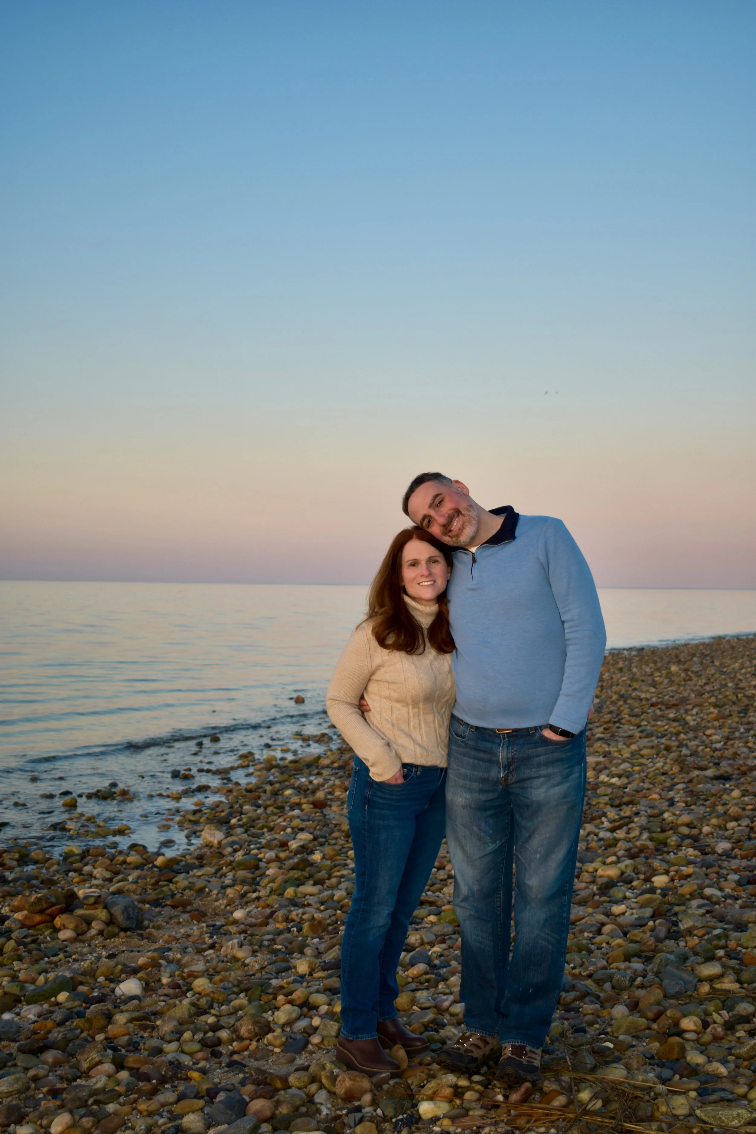 A loving couple standing on a pebble beach at sunset, smiling and embracing near the water. Ready to adopt a baby.