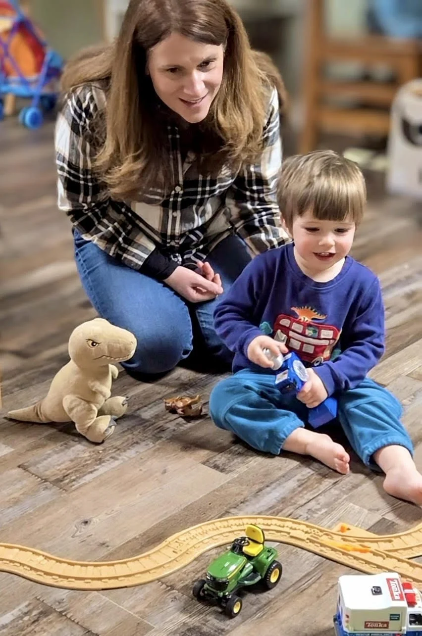 A woman and a young boy sitting on a wooden floor surrounded by toys, including a stuffed dinosaur, a toy train, and a toy tractor. The woman is wearing a plaid shirt and jeans, and the boy is wearing a blue long-sleeve shirt and blue pants, holding a toy.