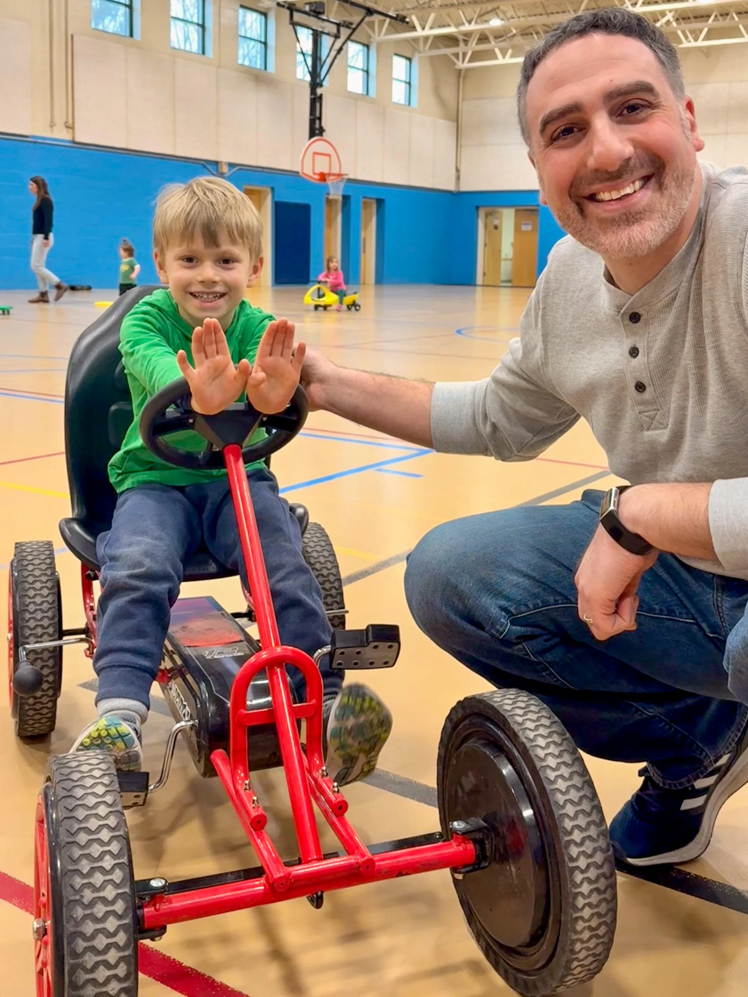 A young boy sitting on a red tricycle inside a gymnasium, smiling and waving at the camera, with a man crouching beside him also smiling. In the background, a blue wall, basketball hoop, and other children playing are visible.