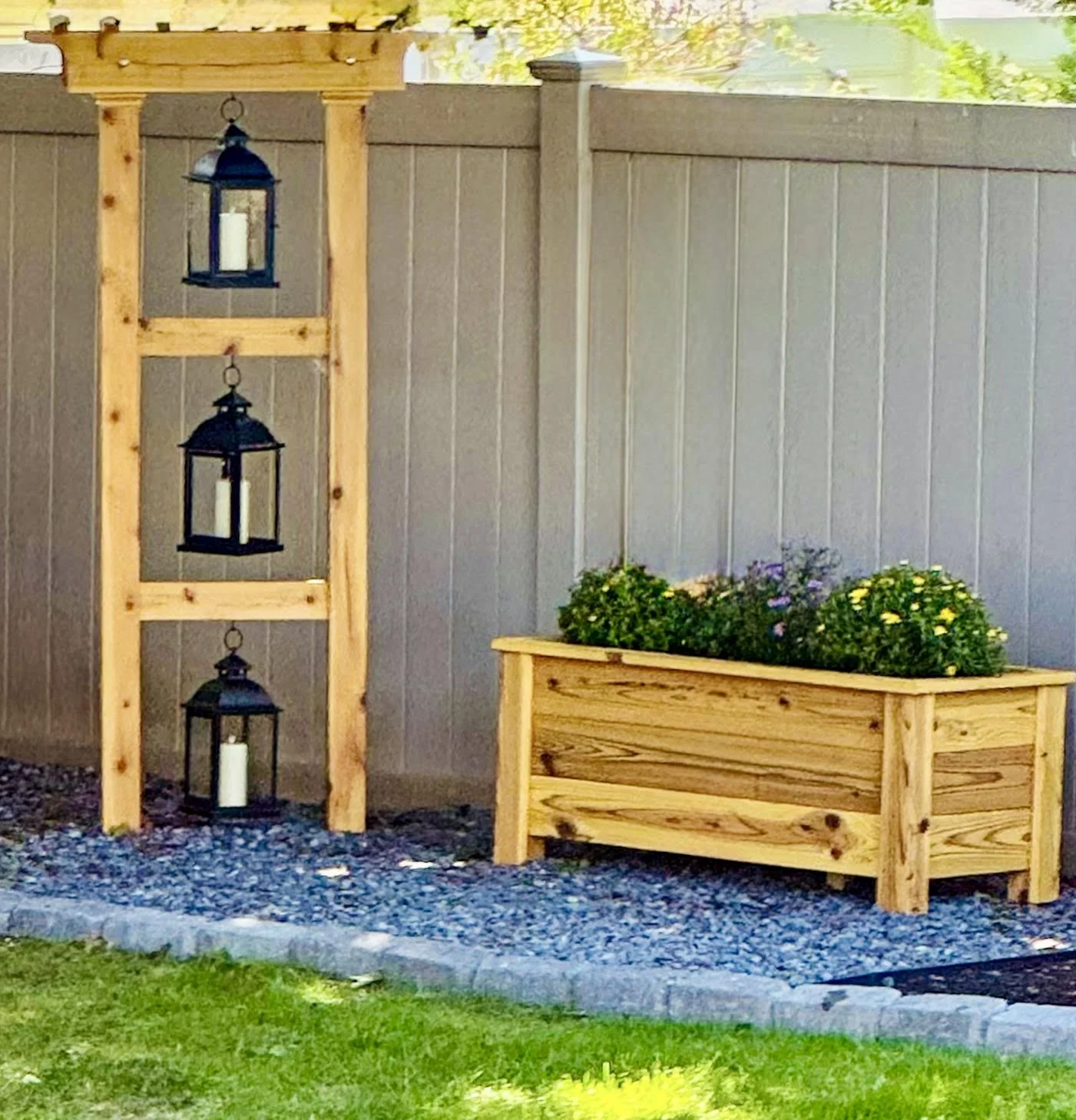 Outdoor garden scene with a wooden planter box filled with flowers and a vertical wooden lantern display with three black lanterns.