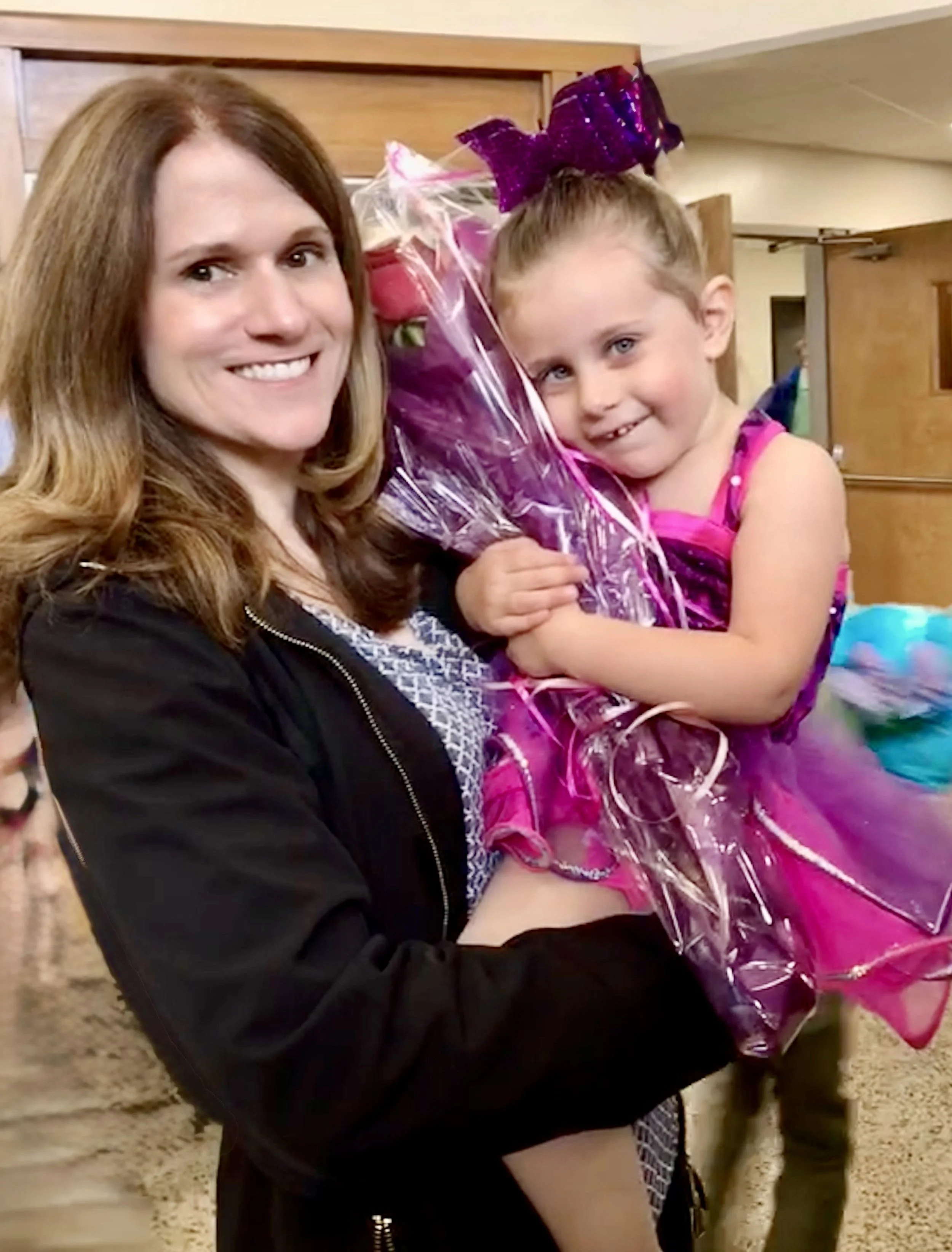 A woman holding a young girl in a colorful dress and large purple bow, both smiling with the woman holding a pink bouquet, in an indoor setting.