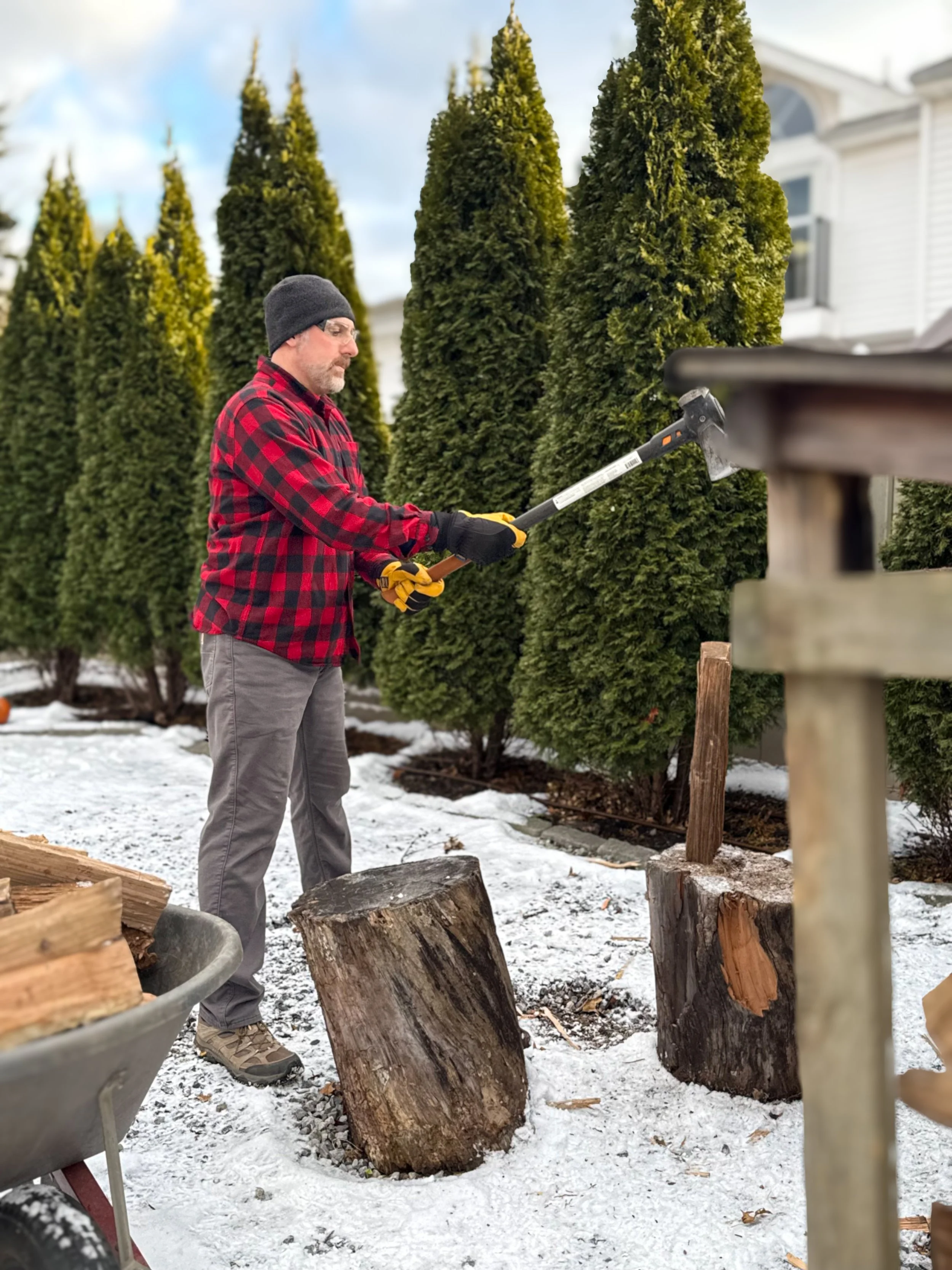 Man in a red and black checkered shirt trimming splitting wood with an ax outdoors in winter, with snow on the ground and tall evergreen trees in the background.