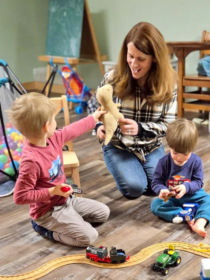 A woman playing with two young boys on a wooden floor, with toy trains, a plush toy, and a ball pit in the background.