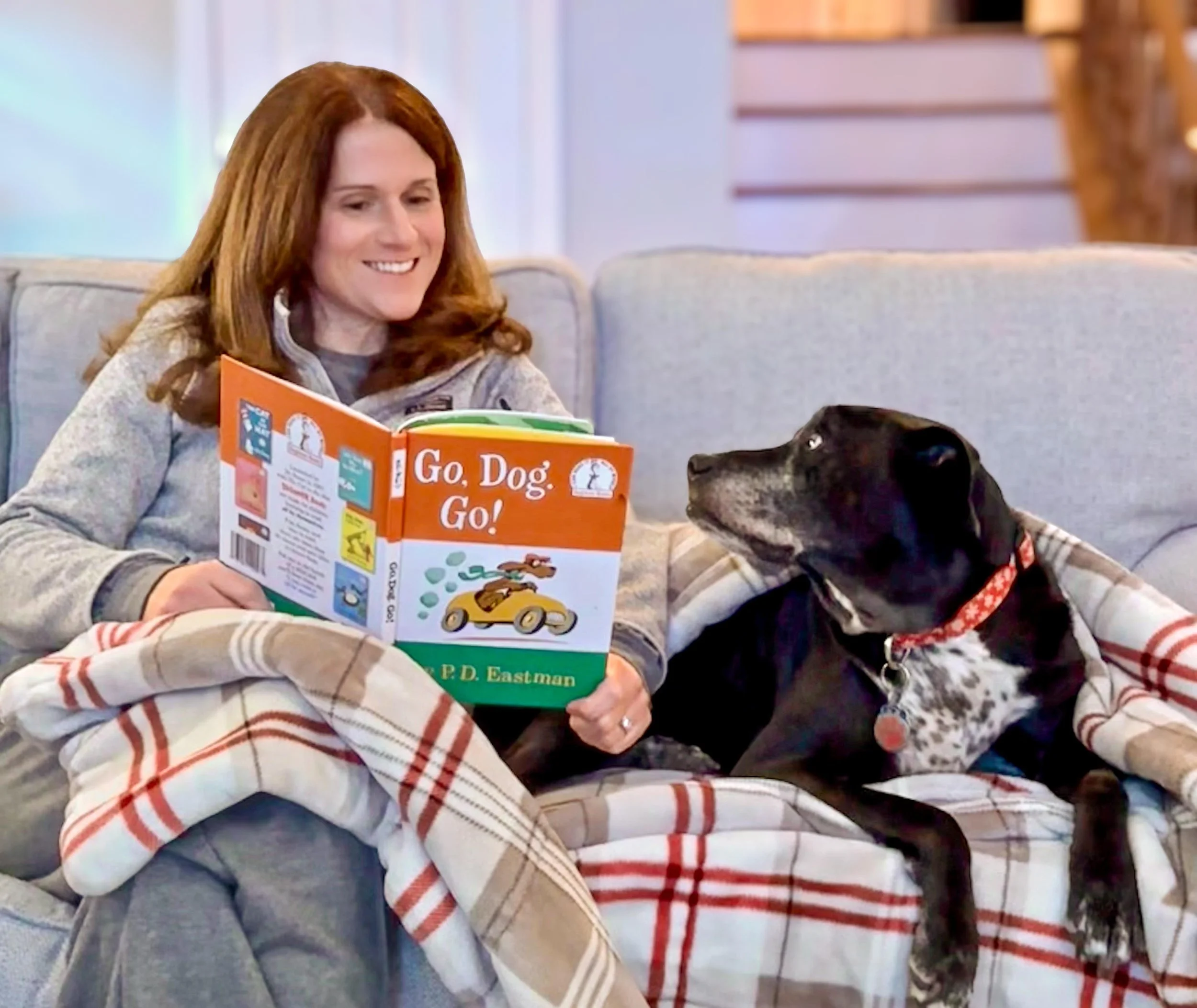 A woman with red hair sitting on a couch, reading a book titled 'Go, Dog. Go!' with a black dog sitting beside her, both covered with a plaid blanket.