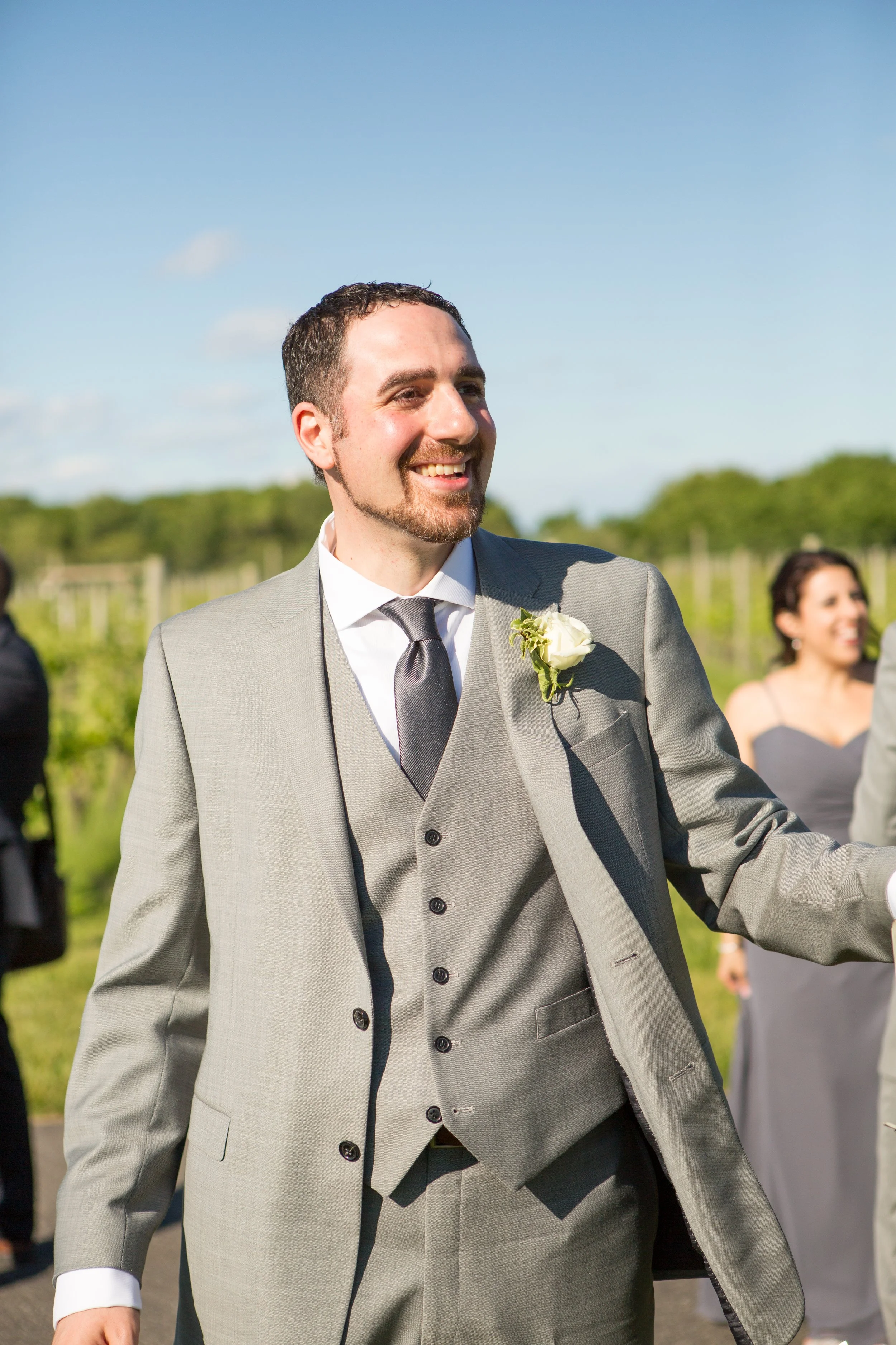 A man in a grey suit and black tie smiling at an outdoor wedding ceremony with a woman in a grey dress in the background.