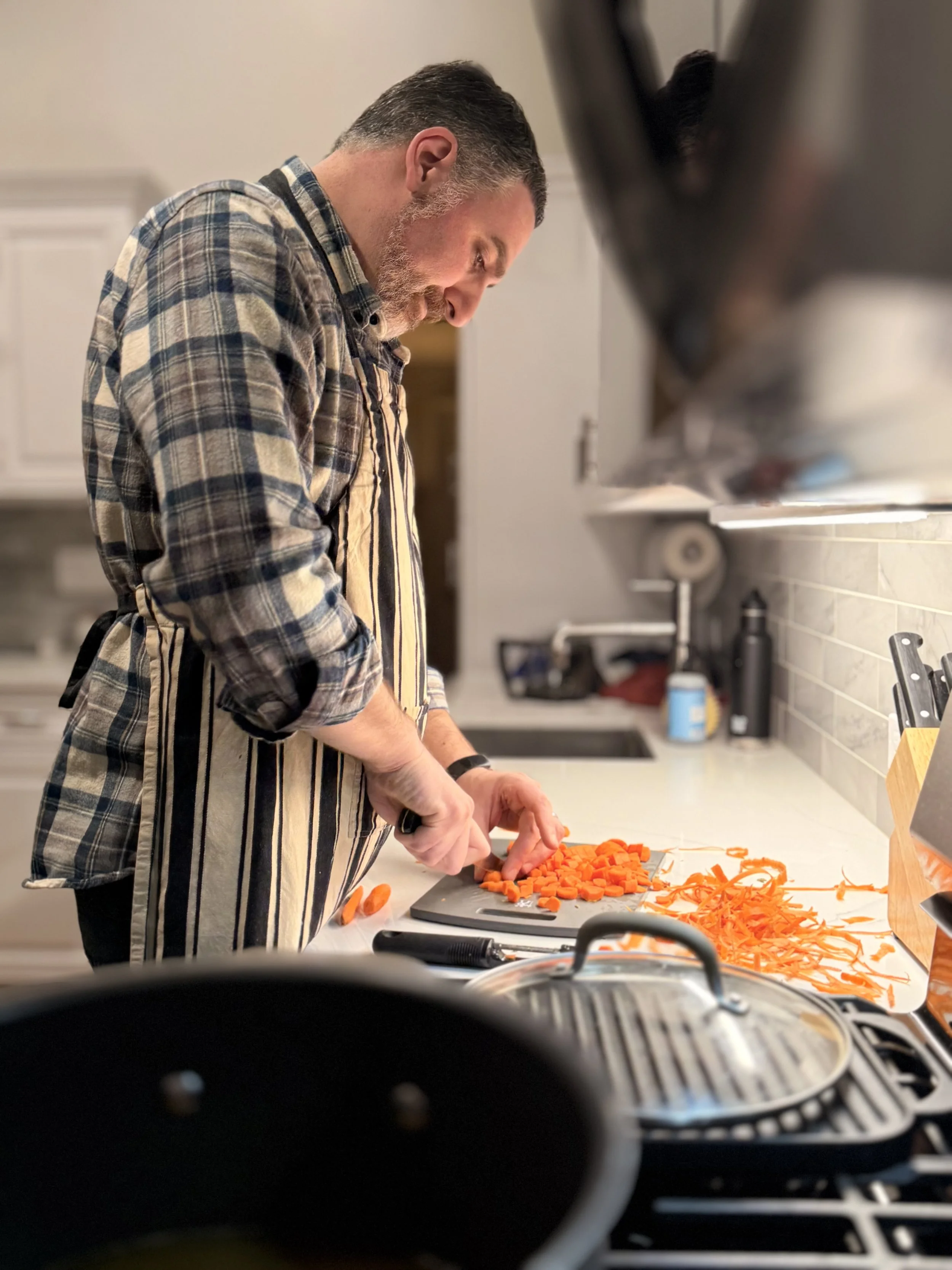 A man with a beard and plaid shirt chopping carrots on a kitchen counter.