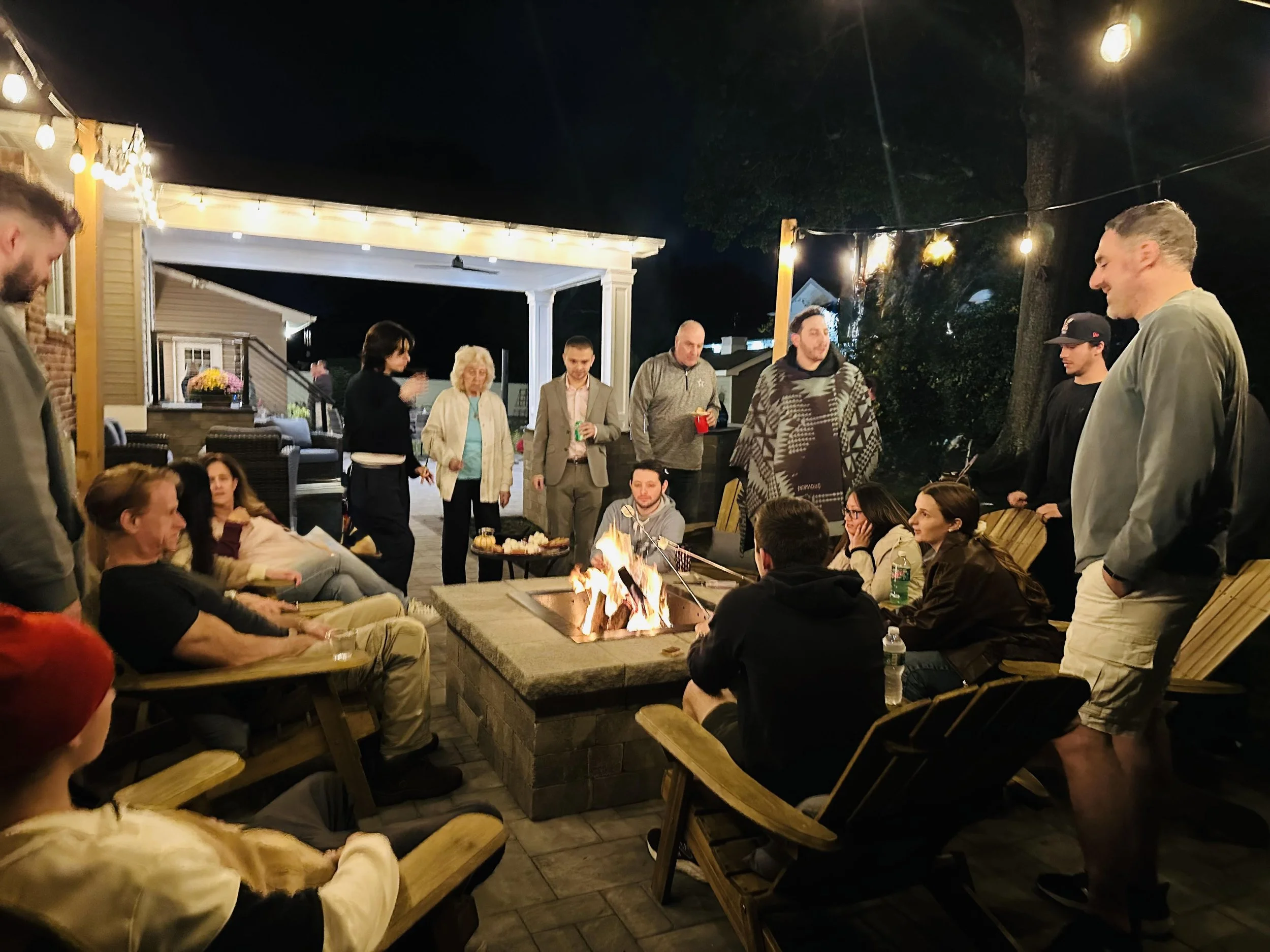 A group of people gathered around a fire pit on a patio during nighttime, some sitting in chairs and others standing, enjoying the evening.