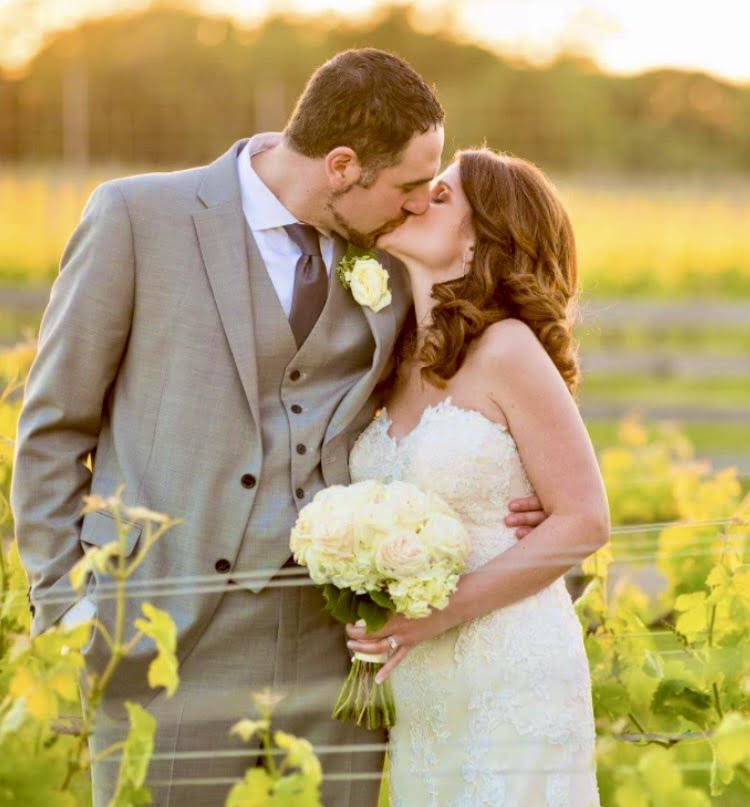 A newlywed couple sharing a kiss in a vineyard during sunset, with the bride holding a bouquet of white flowers and the groom wearing a light gray suit.