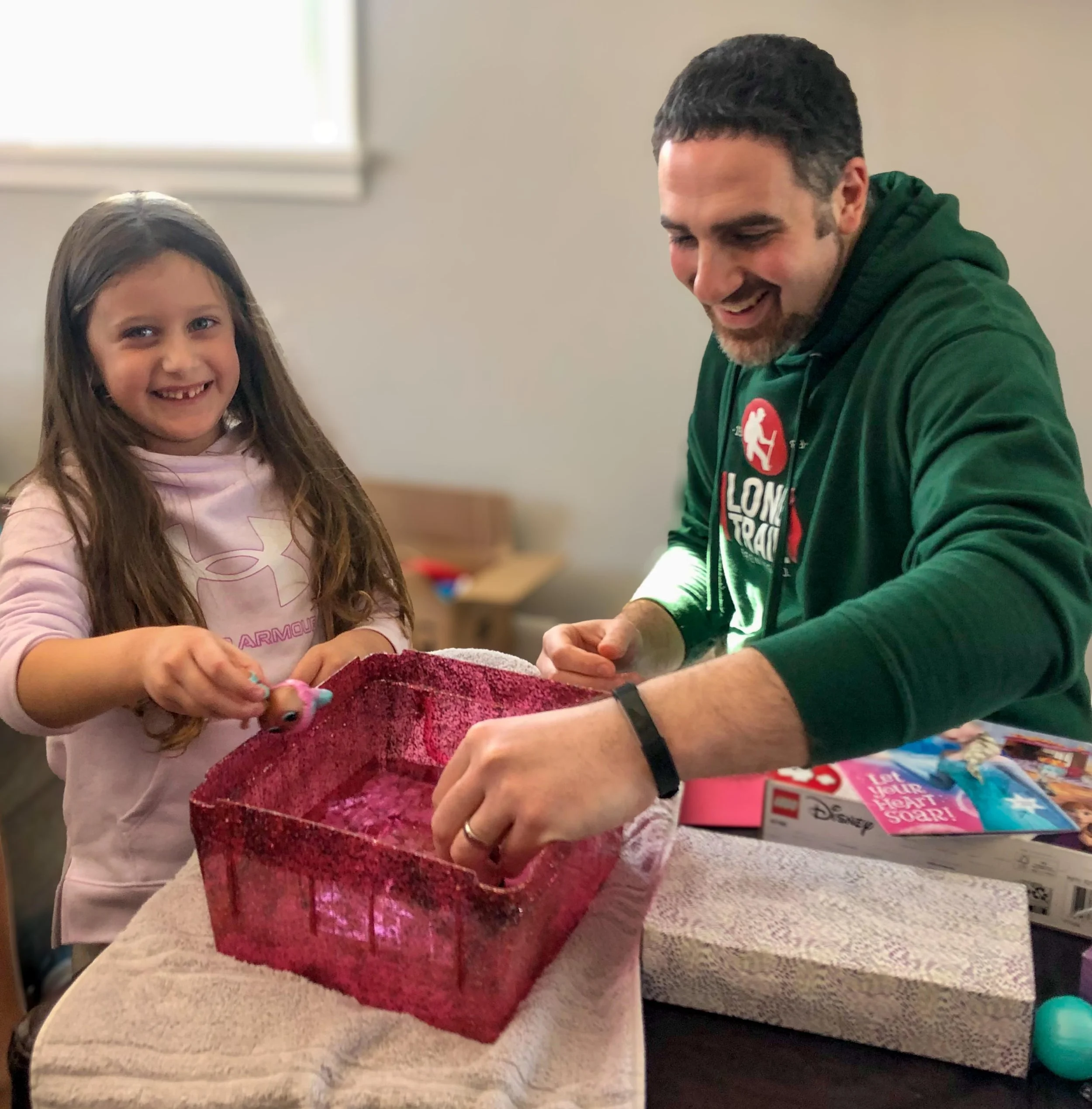 A young girl and a man are happily unboxing a present at a table. The girl is smiling, wearing a pink shirt. The man is smiling, wearing a green hoodie. There is a red box and a Disney puzzle on the table.