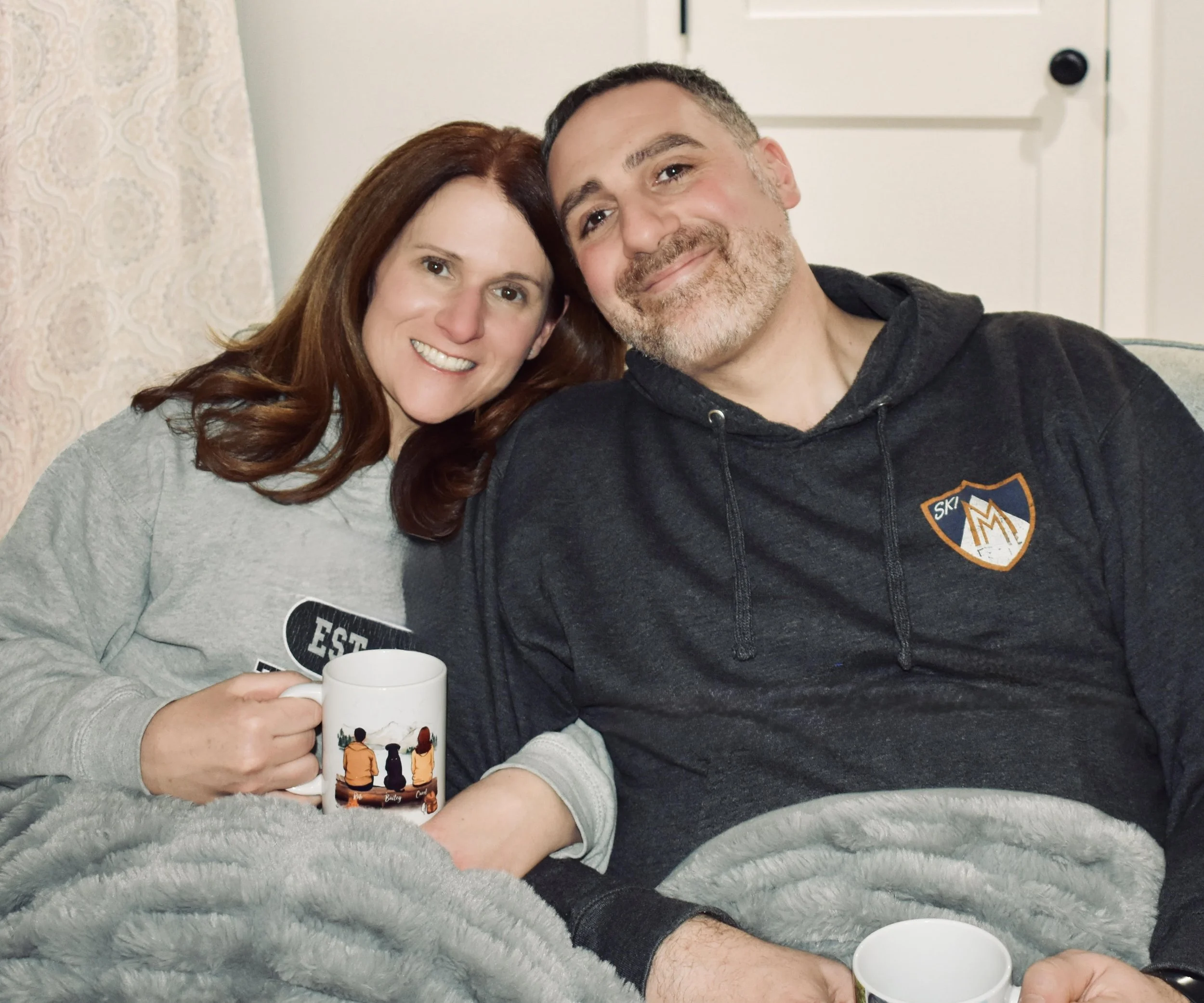 A woman and a man sitting together on a couch, smiling at the camera, holding coffee mugs, wrapped in a gray blanket.