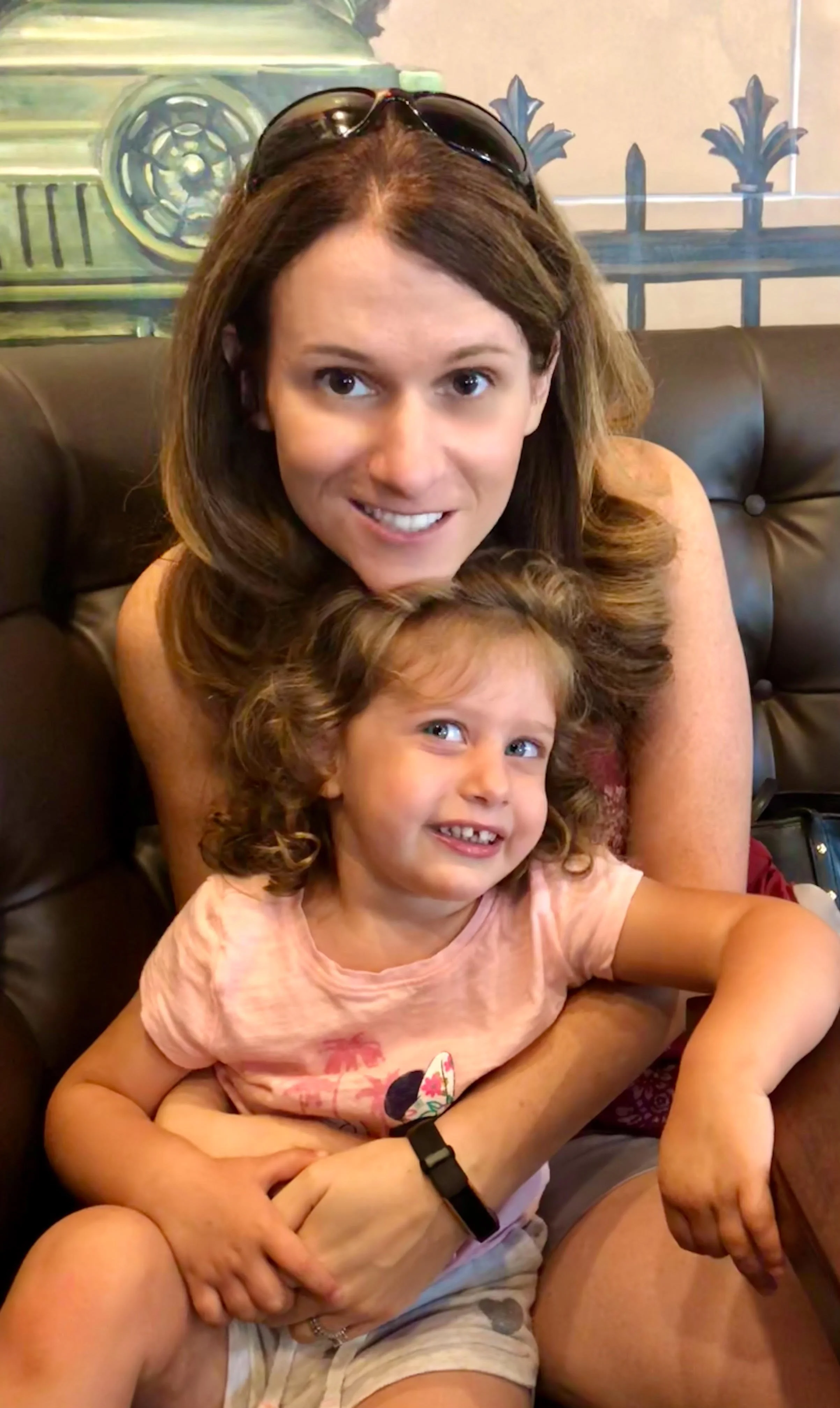 A woman with sunglasses on her head sitting on a leather couch with a young girl in a pink shirt and beige shorts, both smiling.