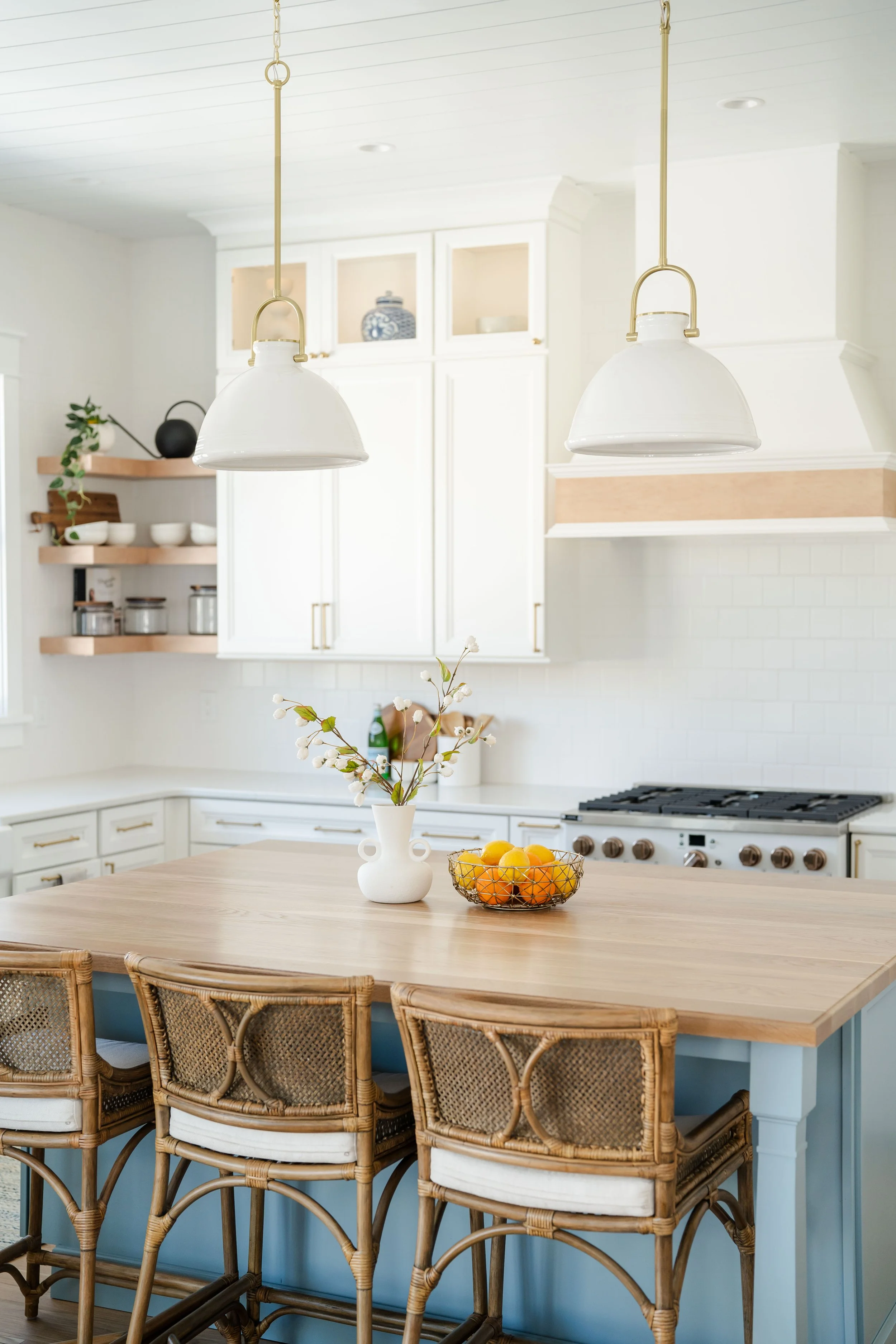 Bright kitchen with white cabinets, a wooden island with three rattan chairs, a vase with flowers, and a bowl of oranges on the island. Two pendant lights hang above the island.