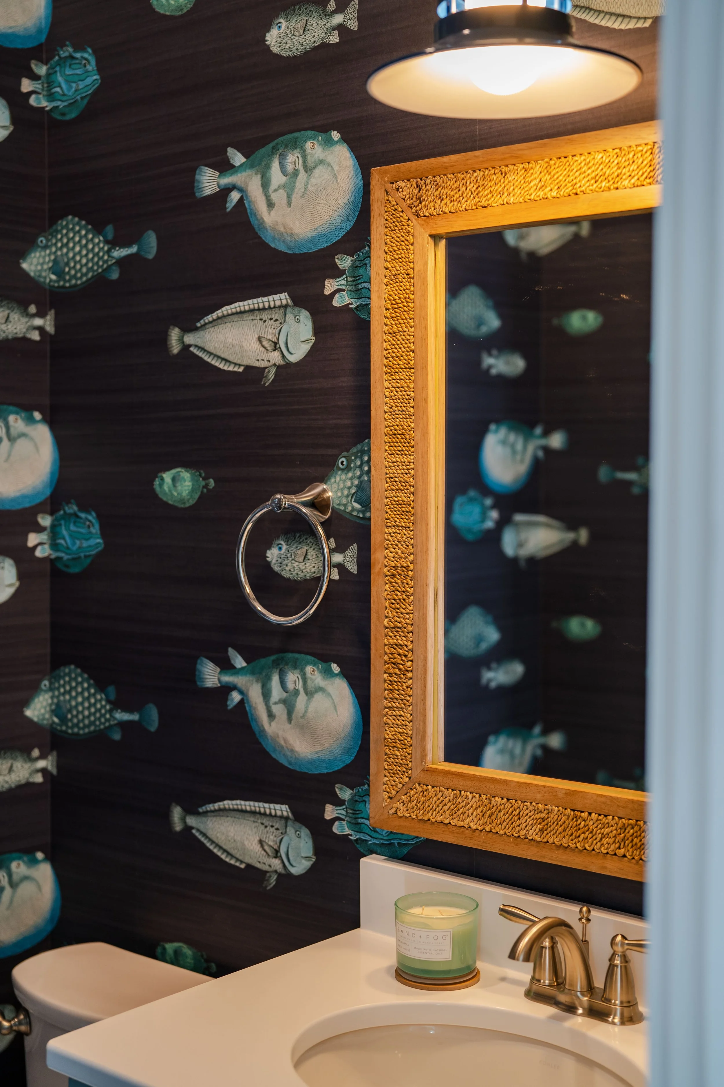 A bathroom with dark wallpaper featuring various blue and gray fish. There is a wooden-framed mirror above a white sink, with a gold-colored soap dispenser and a green candle on the counter. A chrome towel ring is mounted on the wall.