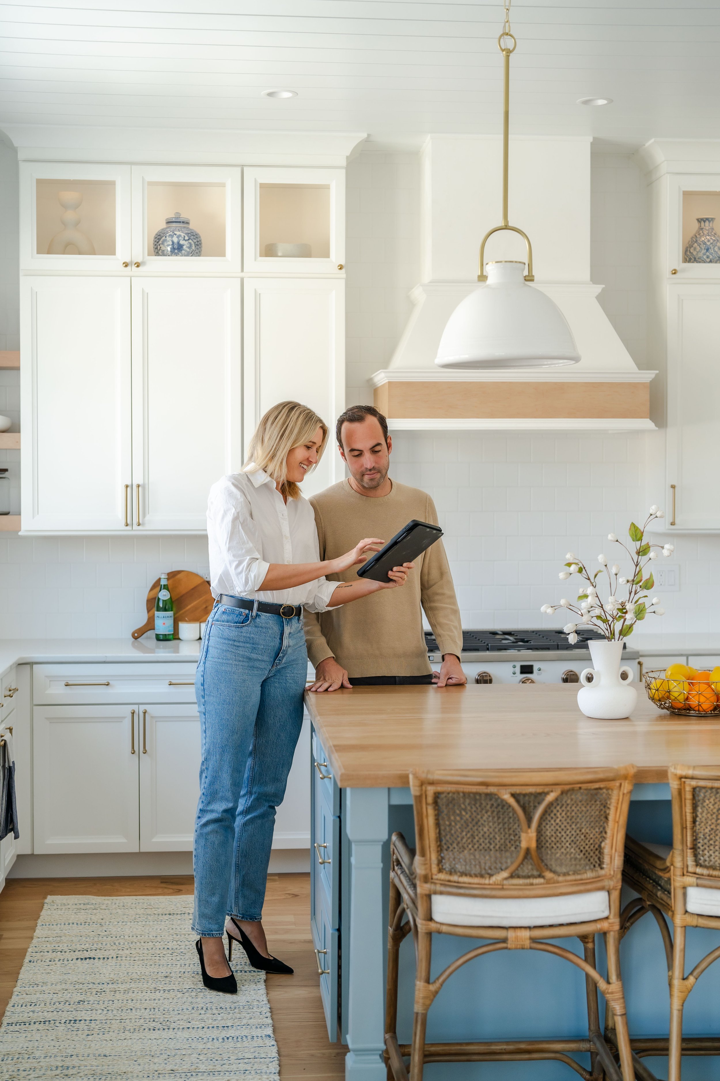 A woman and a man look at a tablet together in a modern kitchen. The woman is wearing blue jeans and black heels, while the man is dressed in a beige sweater. The kitchen has white cabinets, a wooden countertop island with chairs, a white vase with branches, and a fruit bowl with oranges.