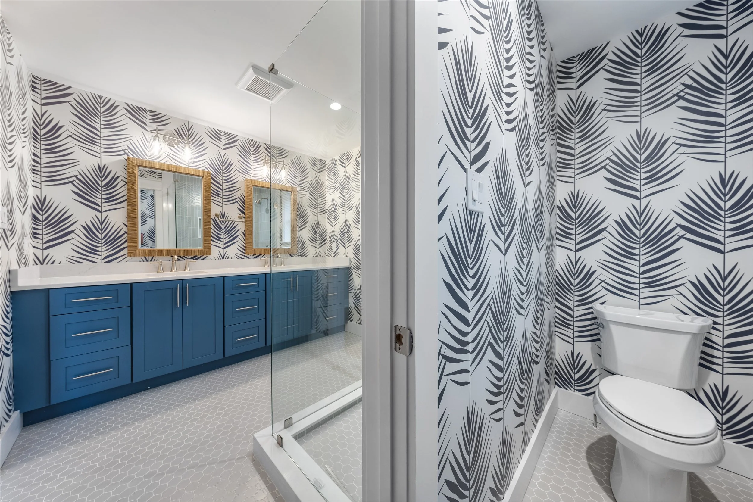 A bathroom with tropical leaf patterned wallpaper, a double vanity with blue cabinets and twin mirrors, a walk-in shower with glass enclosure, and a separate toilet area.