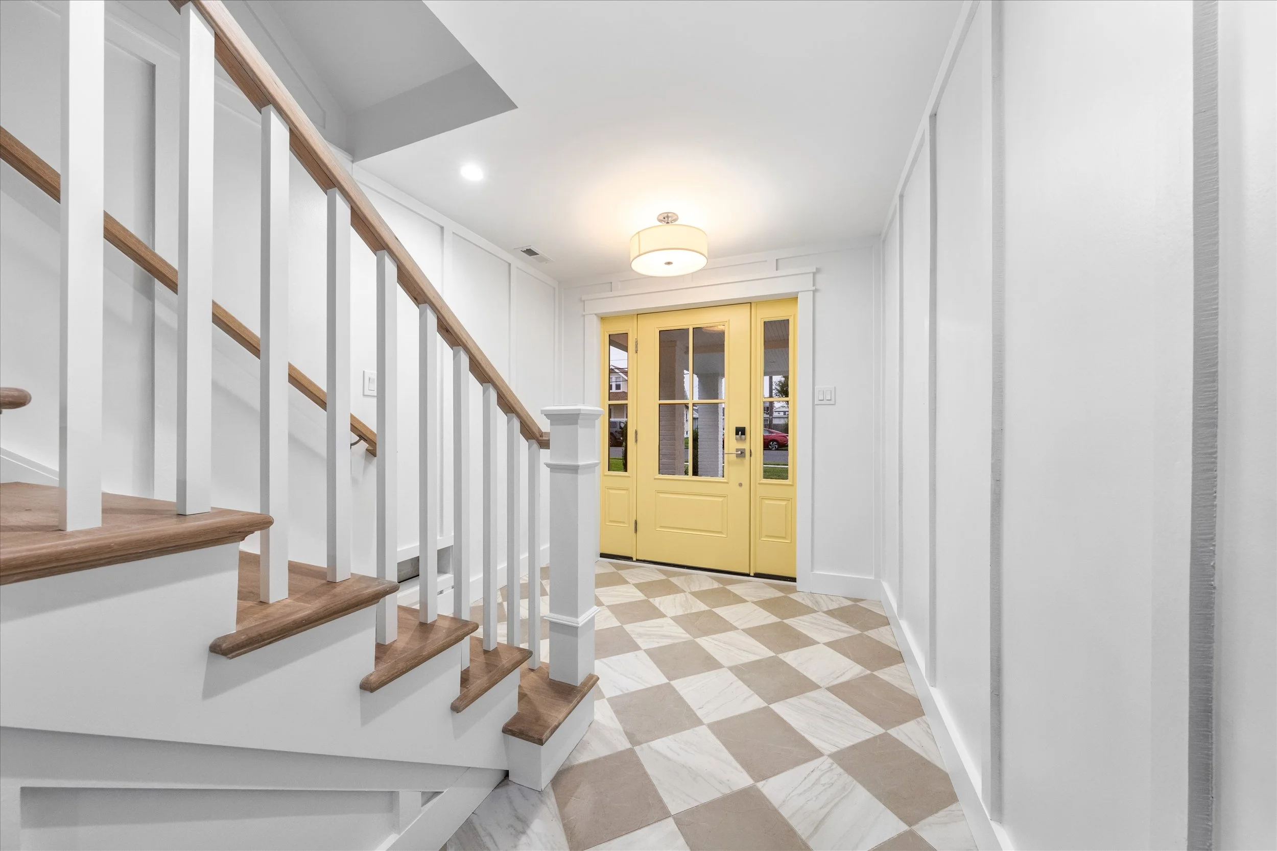 Bright front entryway with yellow door, white walls, checkered patterned tile floor, and staircase with wooden steps and white railing. Built by Vallese builders in North Wildwood, NJ.
