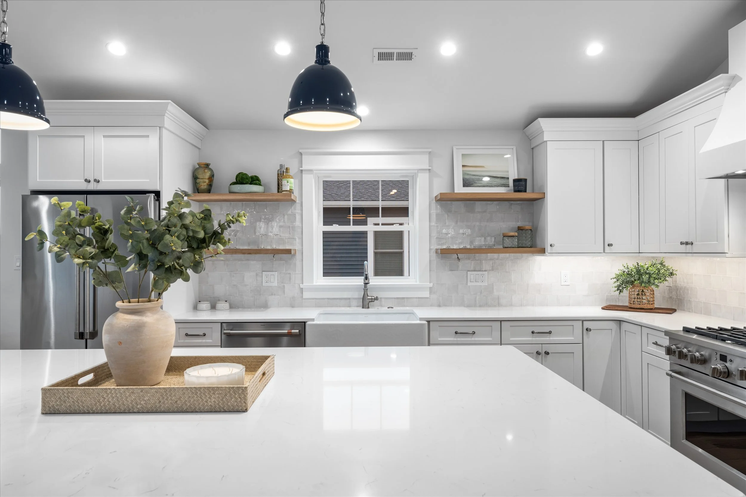 Modern kitchen with white cabinets, marble countertops, black pendant lights, and decorative plants on open wooden shelves.