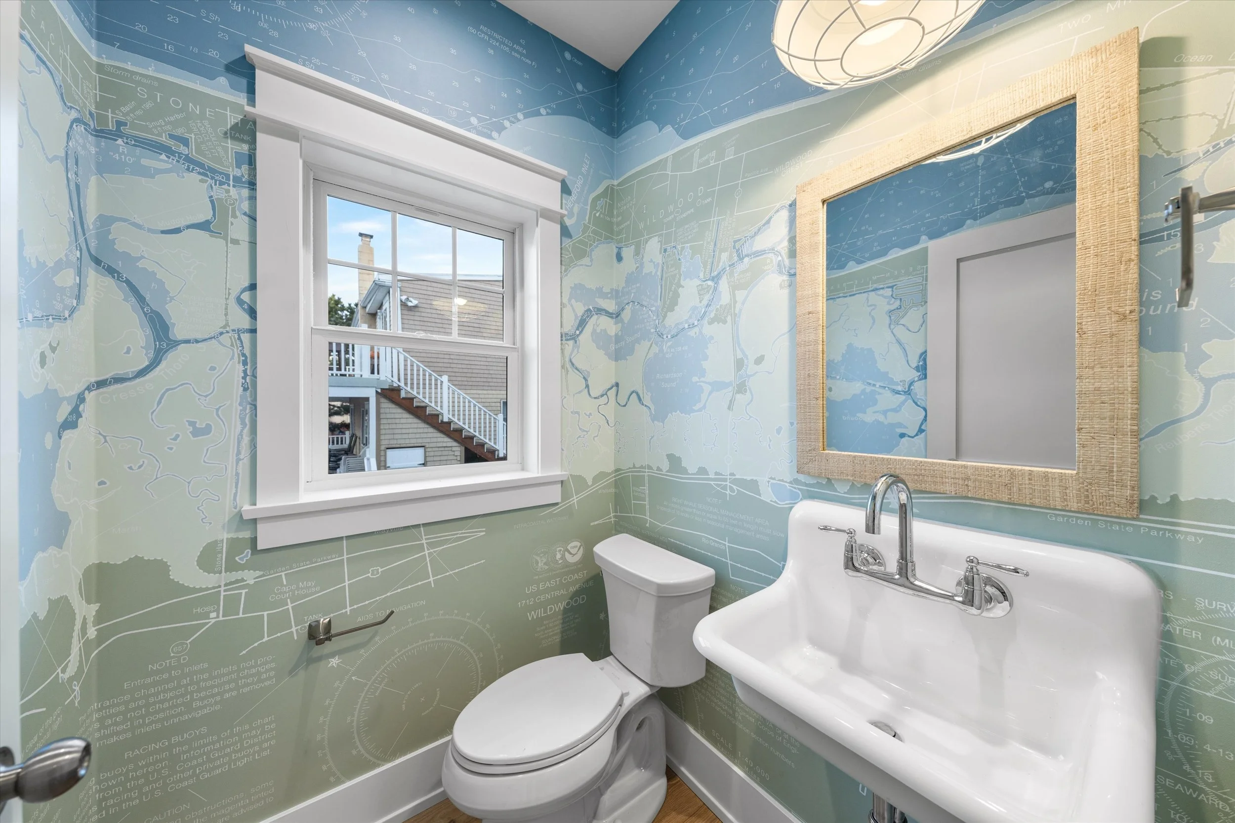 Bathroom with wallpaper featuring nautical charts of north wildwood, a window, a mirror with a textured frame, a toilet, and a sink.