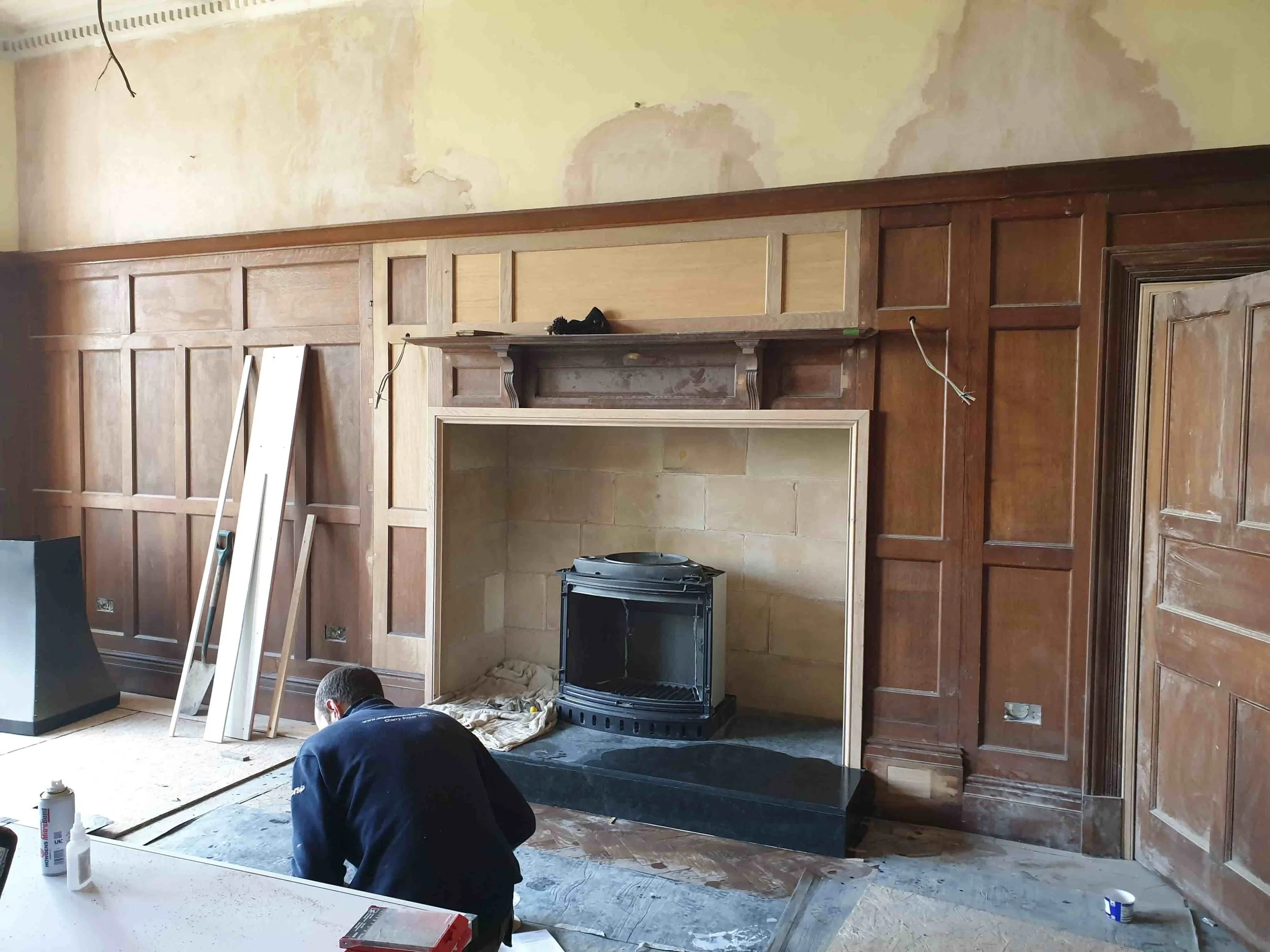 A man working on a fireplace surround installation in a room with wooden wall paneling. Construction tools and materials are scattered around.