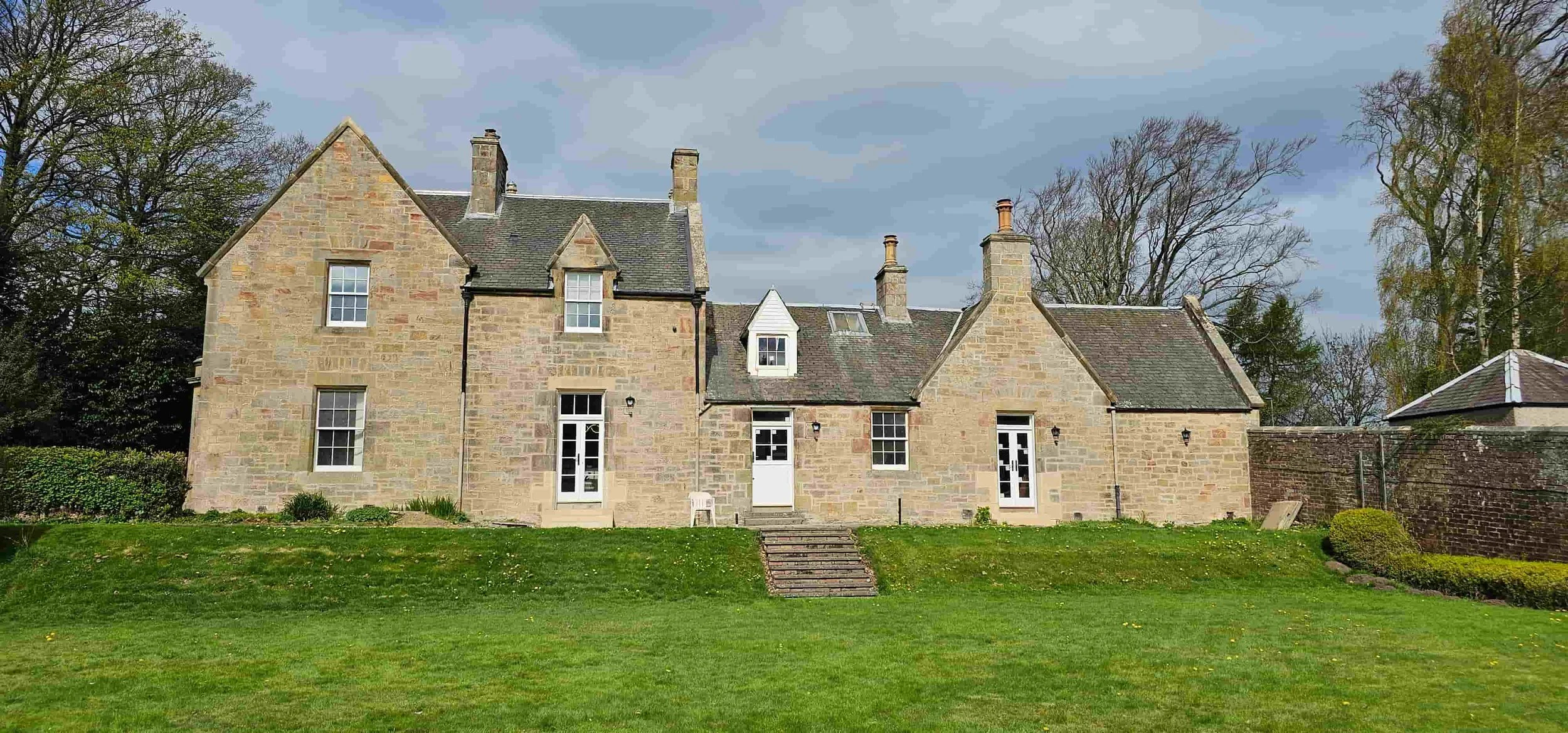 Stone house with multiple chimneys, set on a grassy lawn with trees in the background.