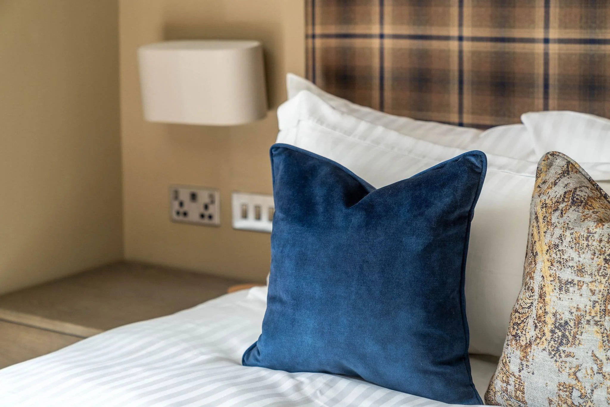 Decorative pillows on a neatly made bed in a hotel room, with a wooden headboard and bedside lamp.