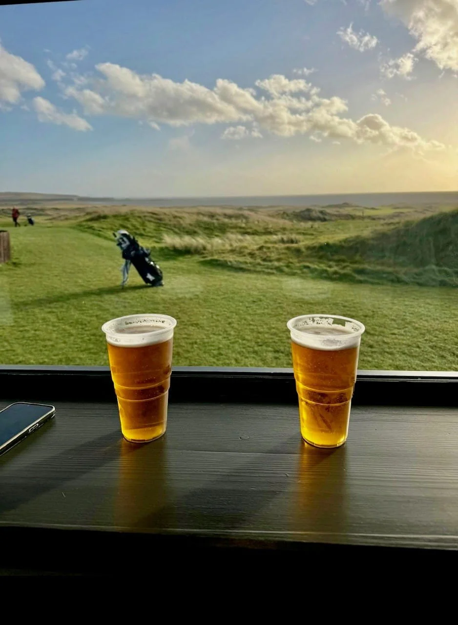Two glasses of beer on a dark table with a window showing a grassy golf course and cloudy sky outside.