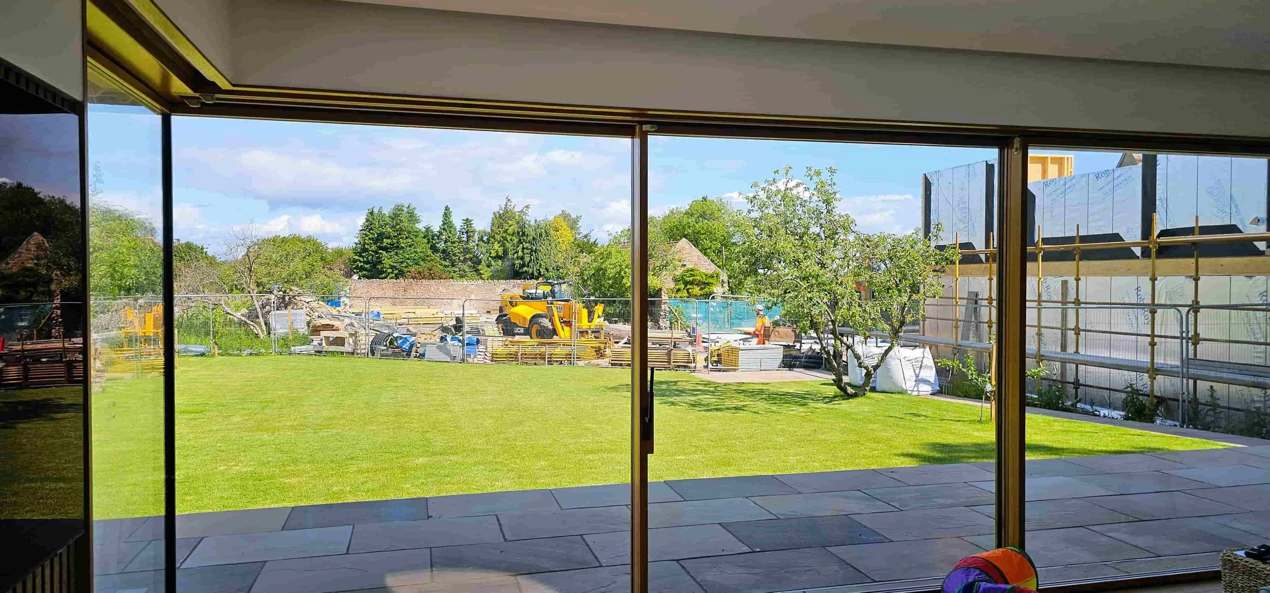 View from inside a house looking out through sliding glass doors onto a backyard with a lawn, trees, and construction work in progress with scaffolding, a yellow construction vehicle, and workers.