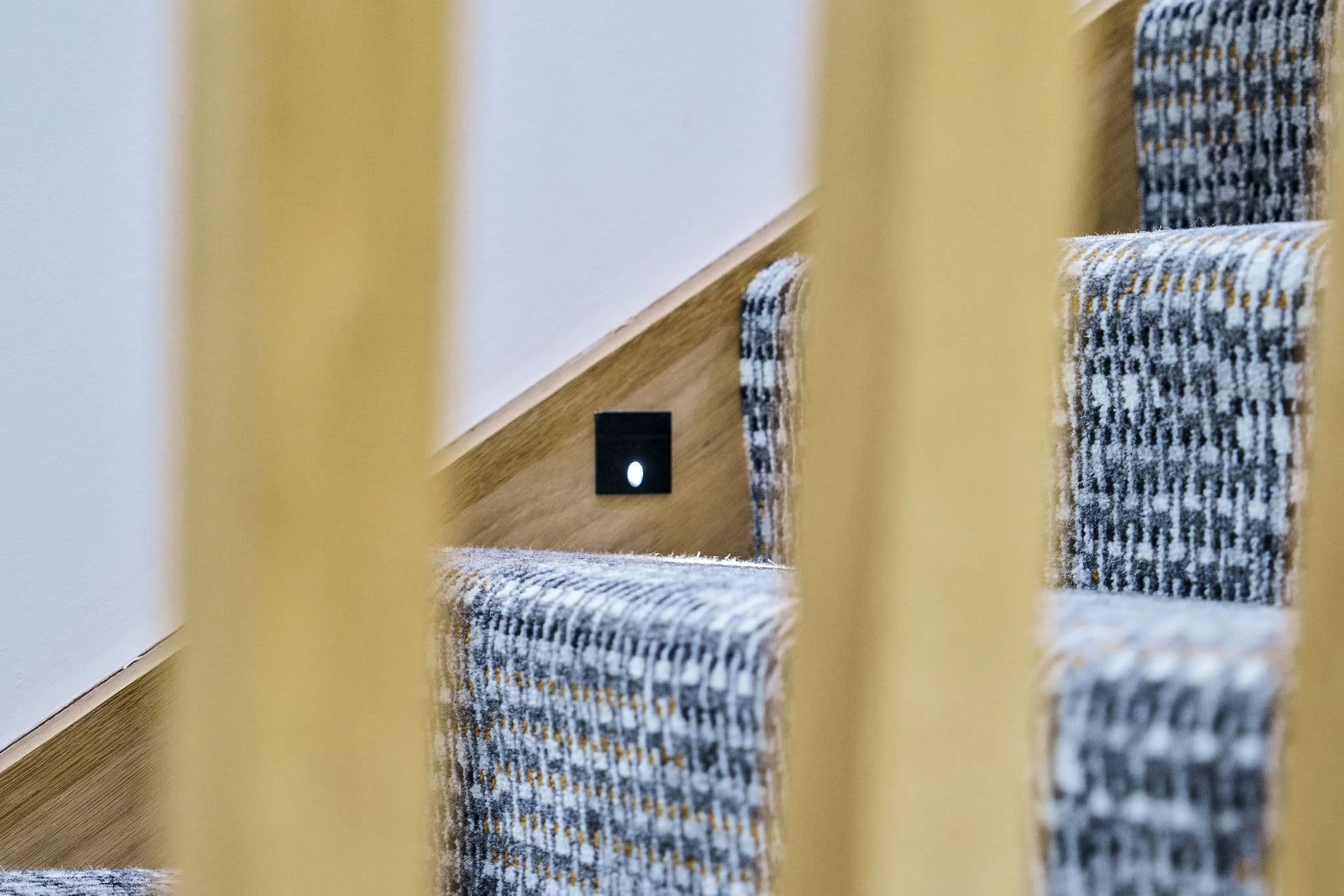 A staircase viewed through vertical railing bars, with patterned carpet on the steps and an electrical outlet on the wall near the stairs.