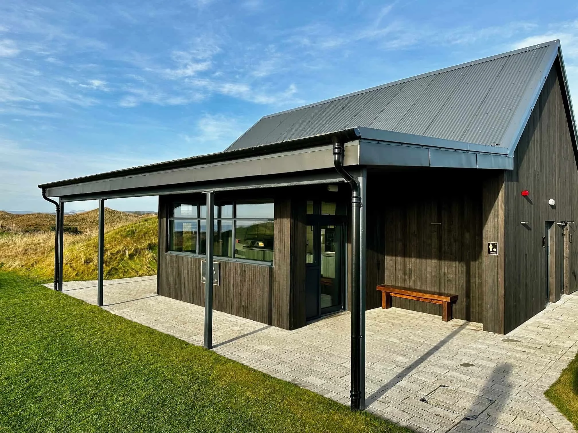 A modern black wooden building with a sloped metal roof, large windows, and a small paved porch with a wooden bench, situated in a grassy landscape under a blue sky.