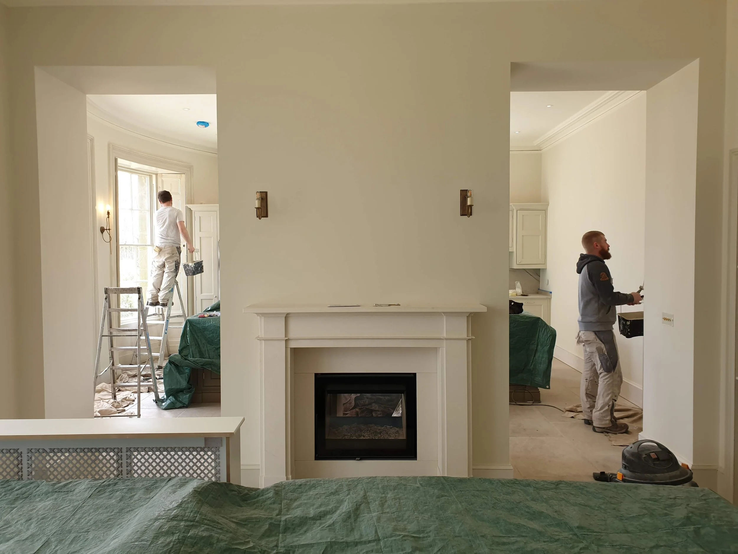 Two workers painting and repairing walls in a home interior. One on a ladder near a window, and the other standing on the floor with tools in hand.