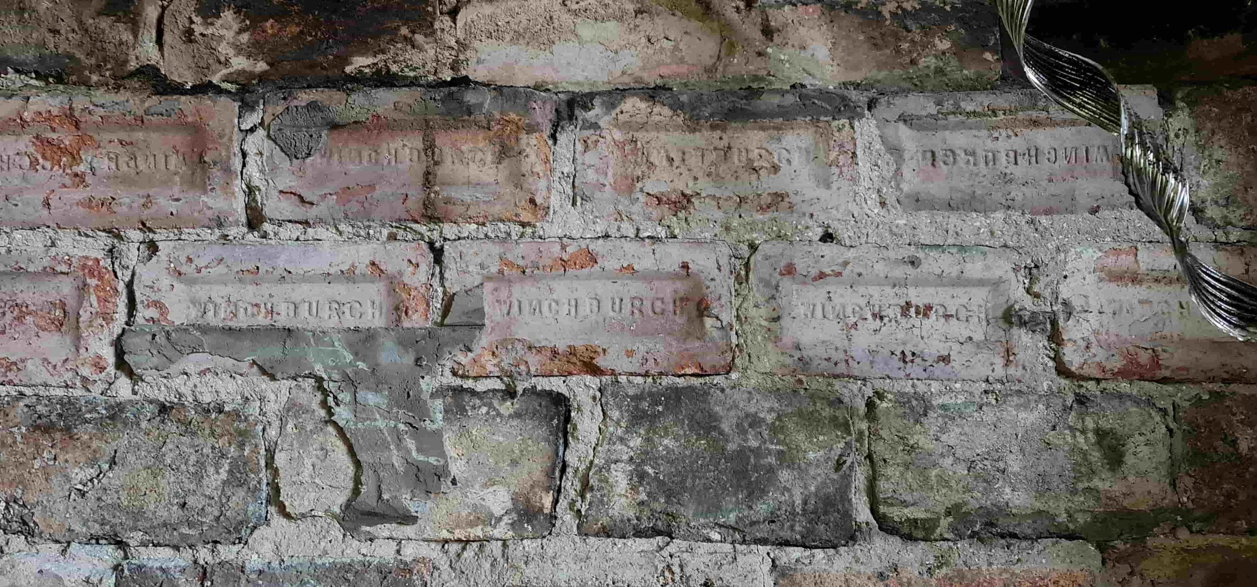 Close-up of an old brick wall with partially faded engraved bricks and some moss or dirt, with a shiny silver ornament partially visible in the upper right corner.
