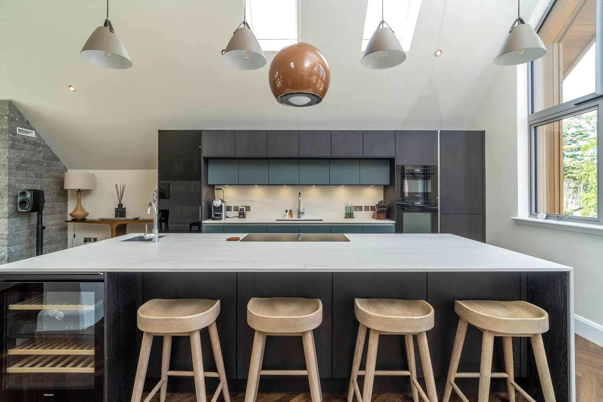 Modern kitchen with black cabinets, white marble countertop island, four wooden stools, and large window with outdoor greenery.