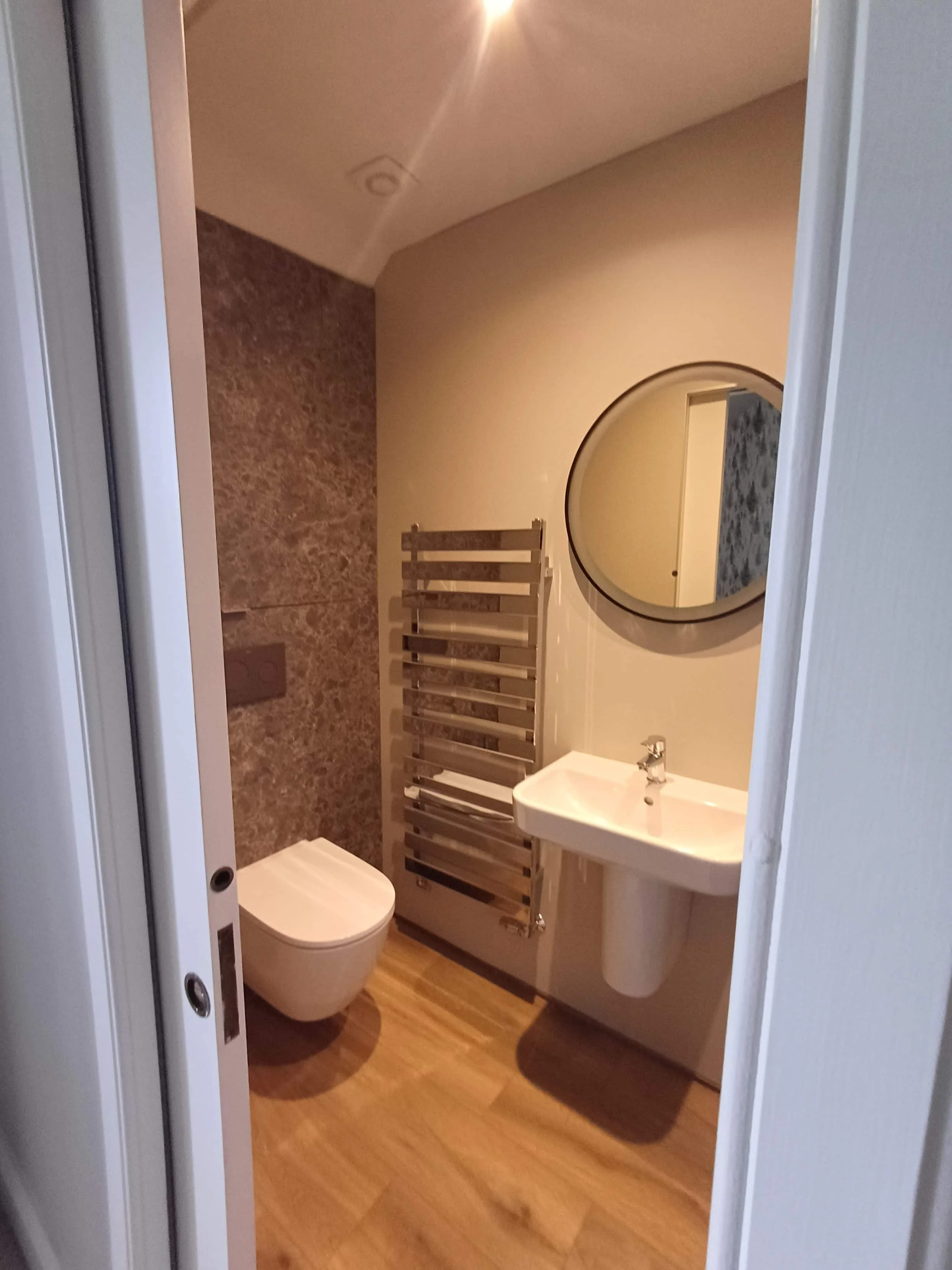 A small bathroom with a wood-look floor, a white wall-mounted sink with a round mirror above , a wall-mounted toilet, and a chrome heated towel rack. The wall behind the toilet features textured brown tiles.