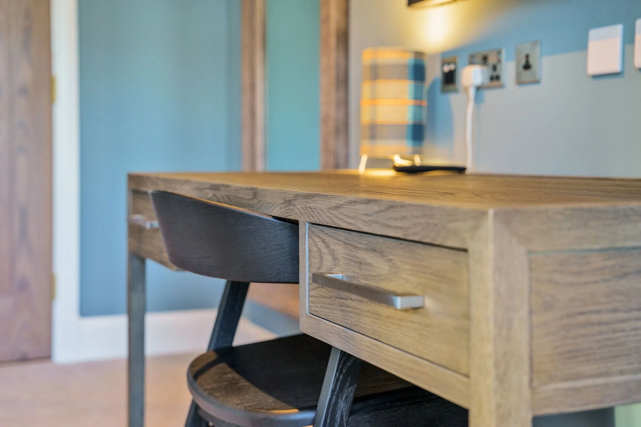 A wooden desk with a drawer and a black chair in a room with a blue wall, electrical outlets, and a lamp in the background.